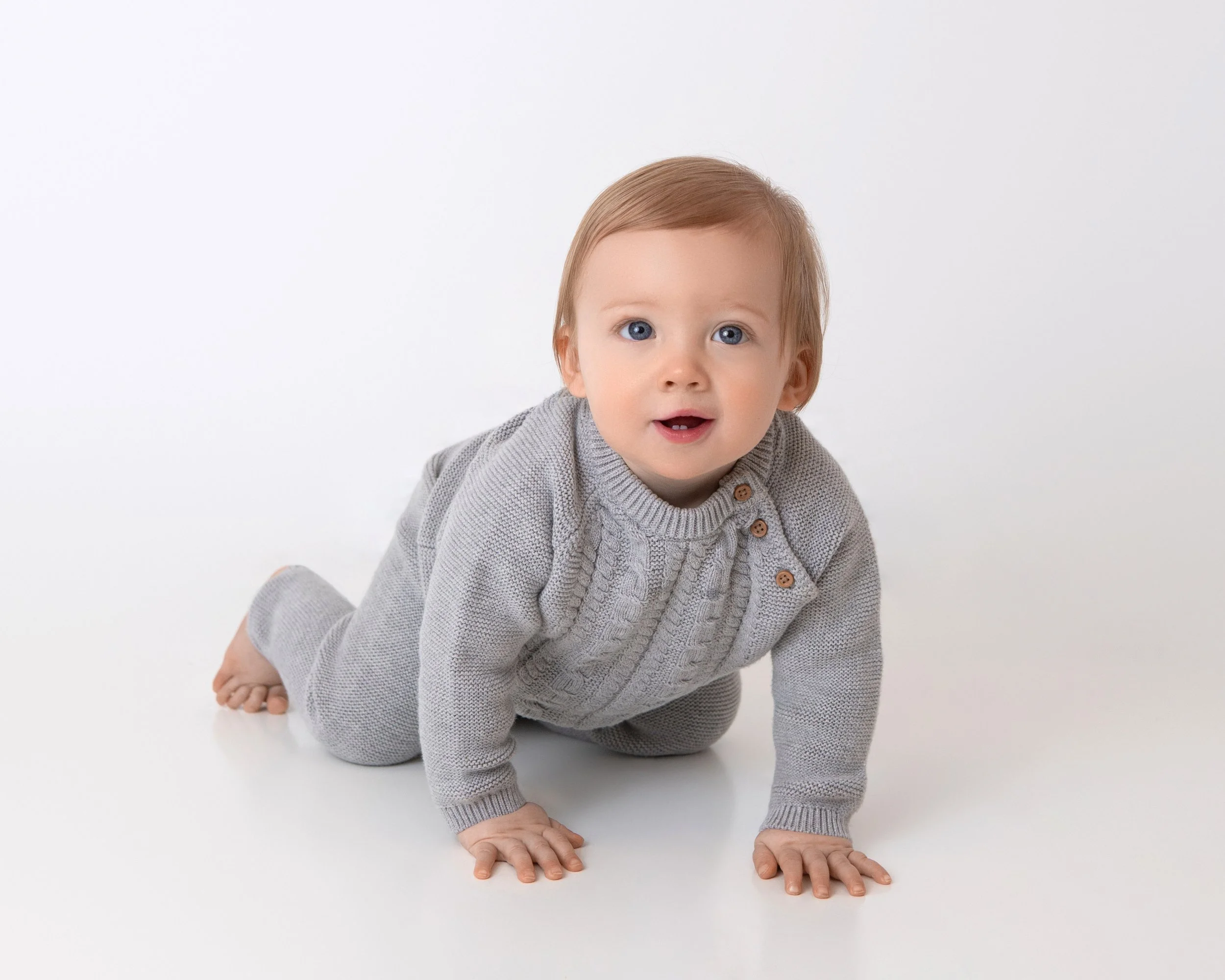 a smiling one year old boy crawling on a white floor wearing a gray colored outfit