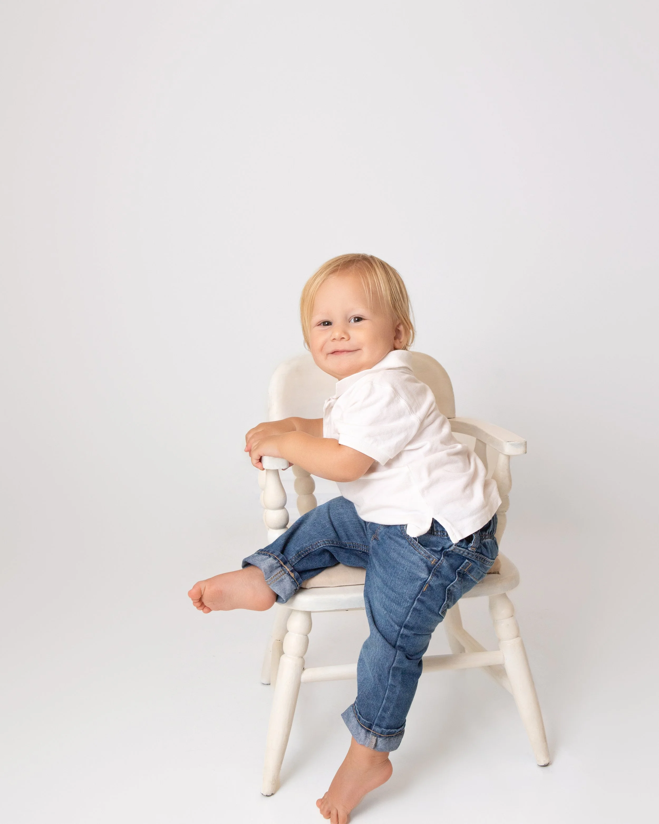 one year old blond boy wearing a white shirt and jeans sitting on a white chair