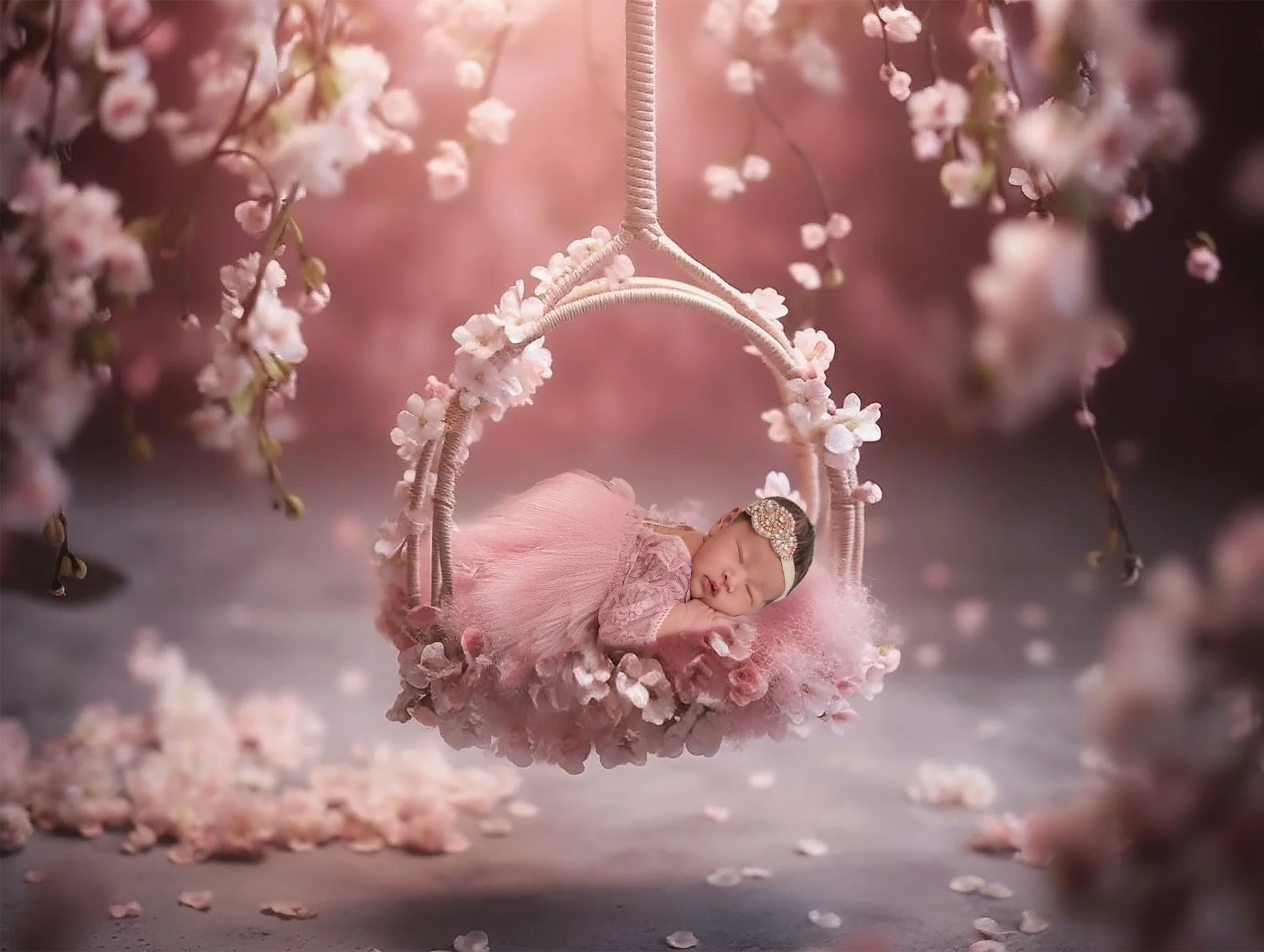 a newborn baby girl in a pink fluffy lace dress sleeping on a swing surrounded by pink flowers and branches