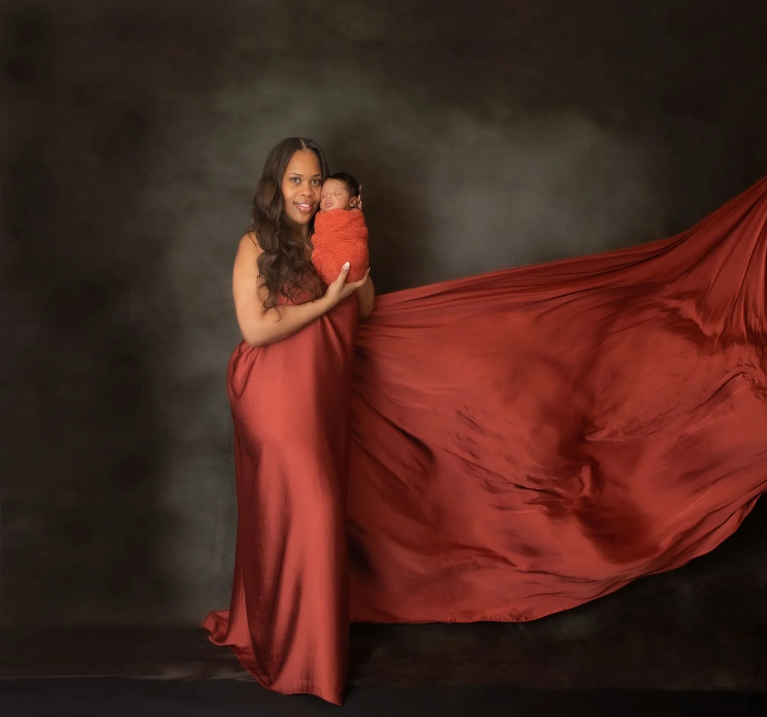 Woman with long dark hair holding a newborn baby wrapped in an orange blanket in front of a dark background, with a long flowing red fabric extending to the right.