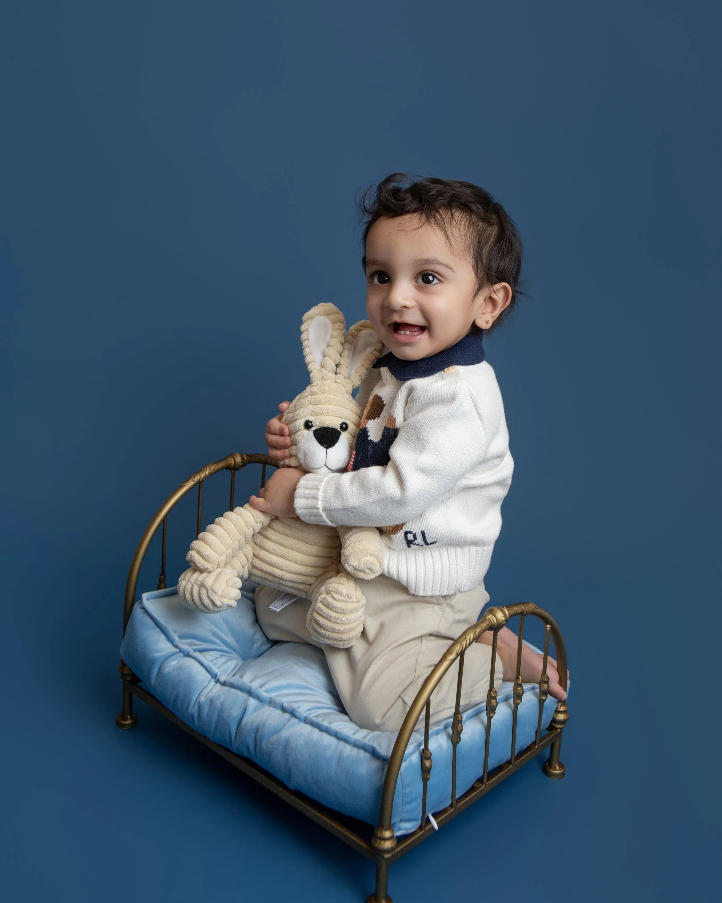 one year old boy smiling holidng a stuffed bunny sitting on a gold bed with blue background