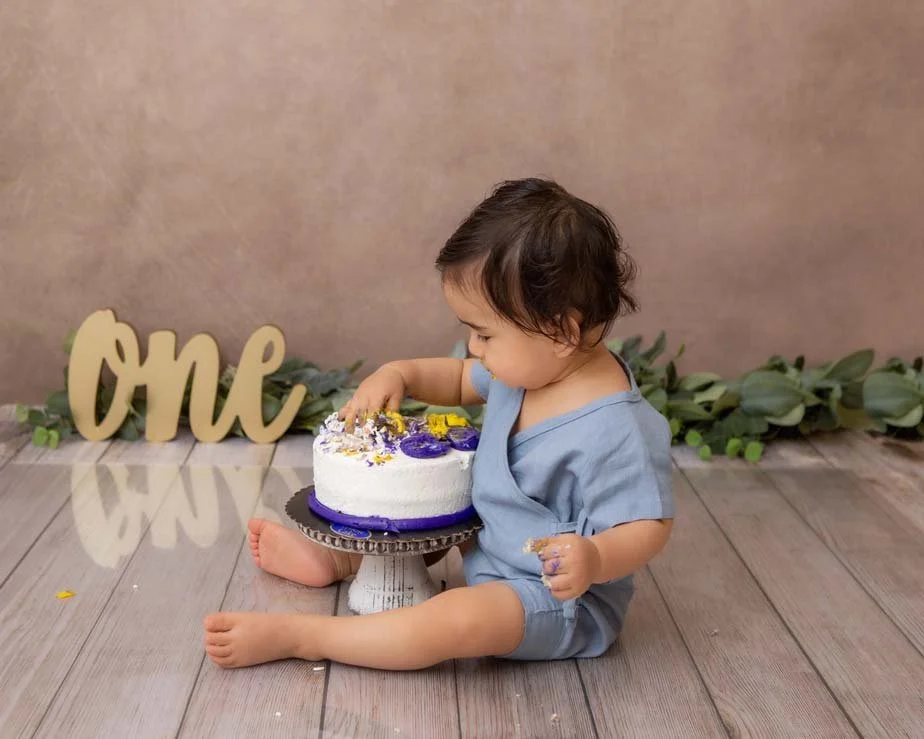 Toddler in a blue romper sitting on a rustic wooden floor, playing and touching a birthday cake decorated with purple and yellow flowers, with a 'One' sign in the background. Simple and sweet first birthday photography in Lancaster, PA.