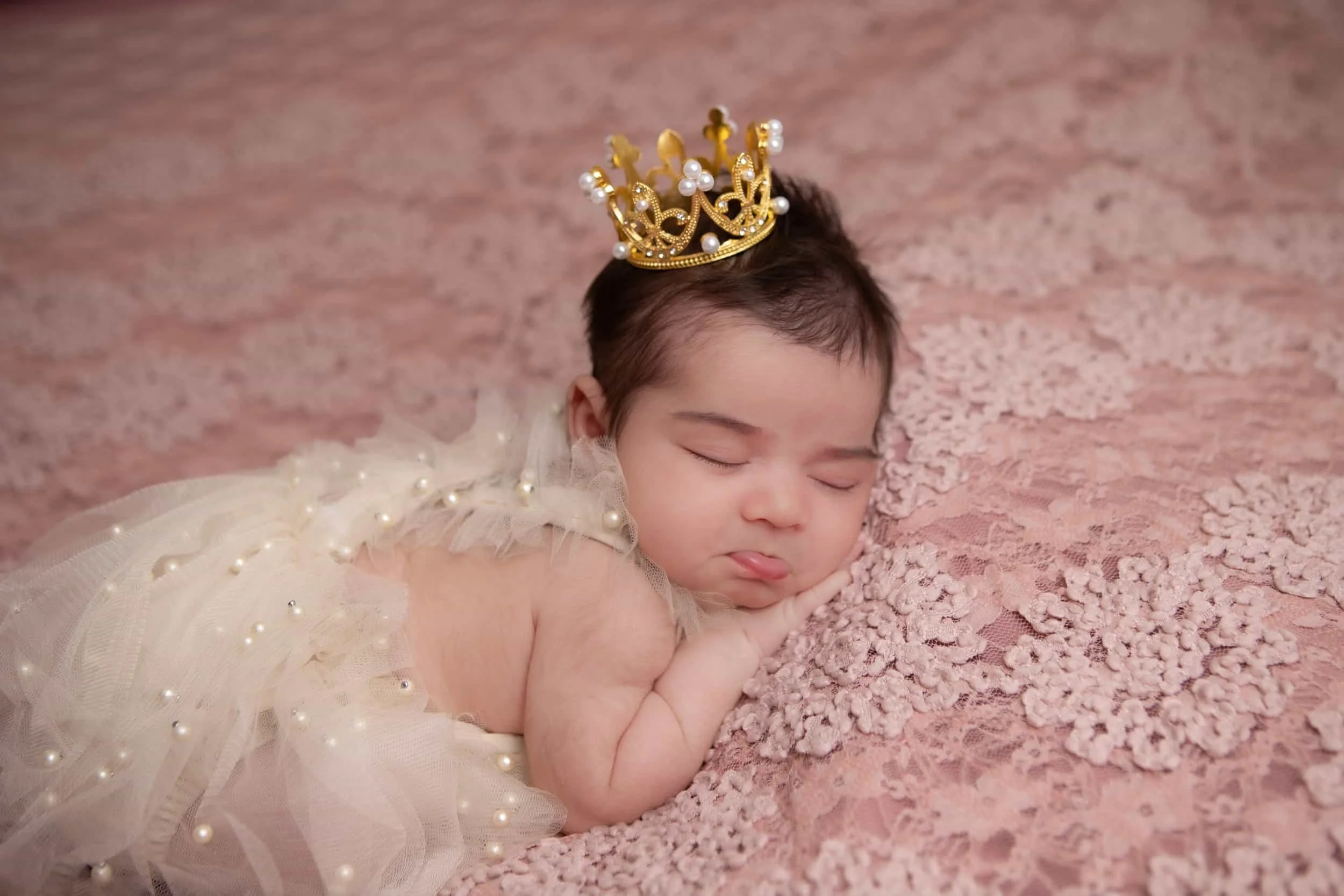 Sleeping newborn baby dressed in a pearl-adorned outfit, wearing a golden crown, resting on a soft lace blanket. Regal and enchanting newborn photography in Lancaster, PA.