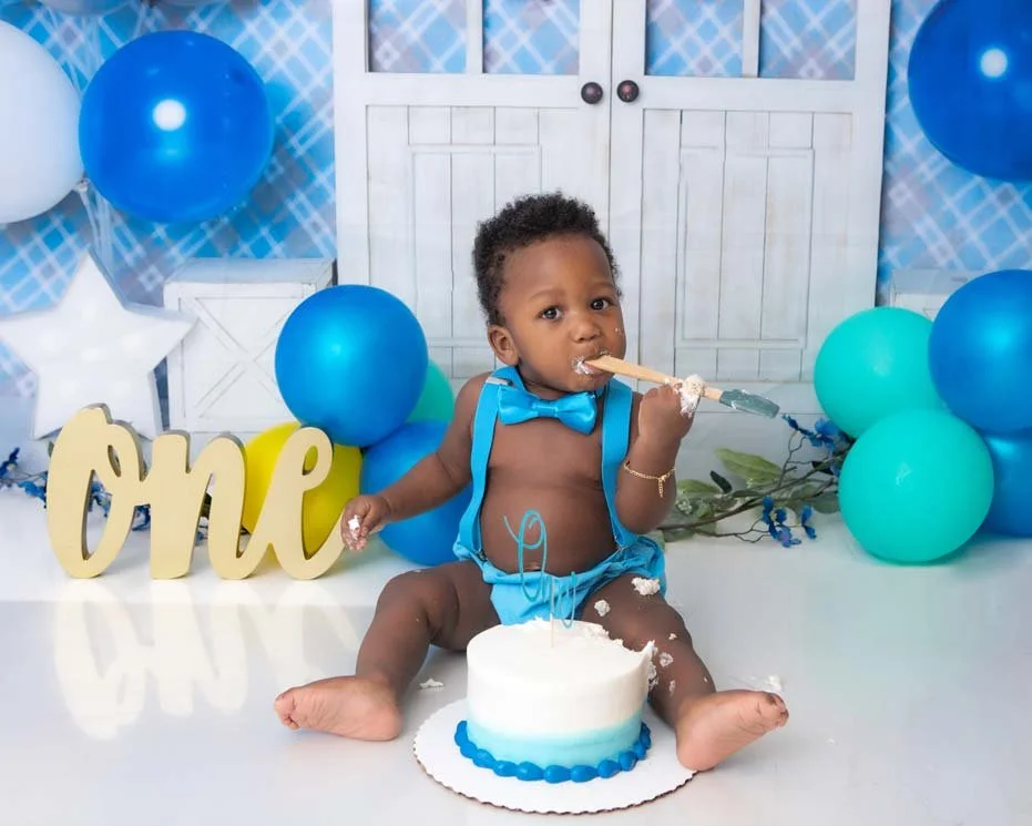Baby boy in a blue bow tie and suspenders, joyfully digging into his birthday cake with a wooden spoon, surrounded by blue balloons and a 'One' sign. Fun and festive cake smash photography in Lancaster, PA