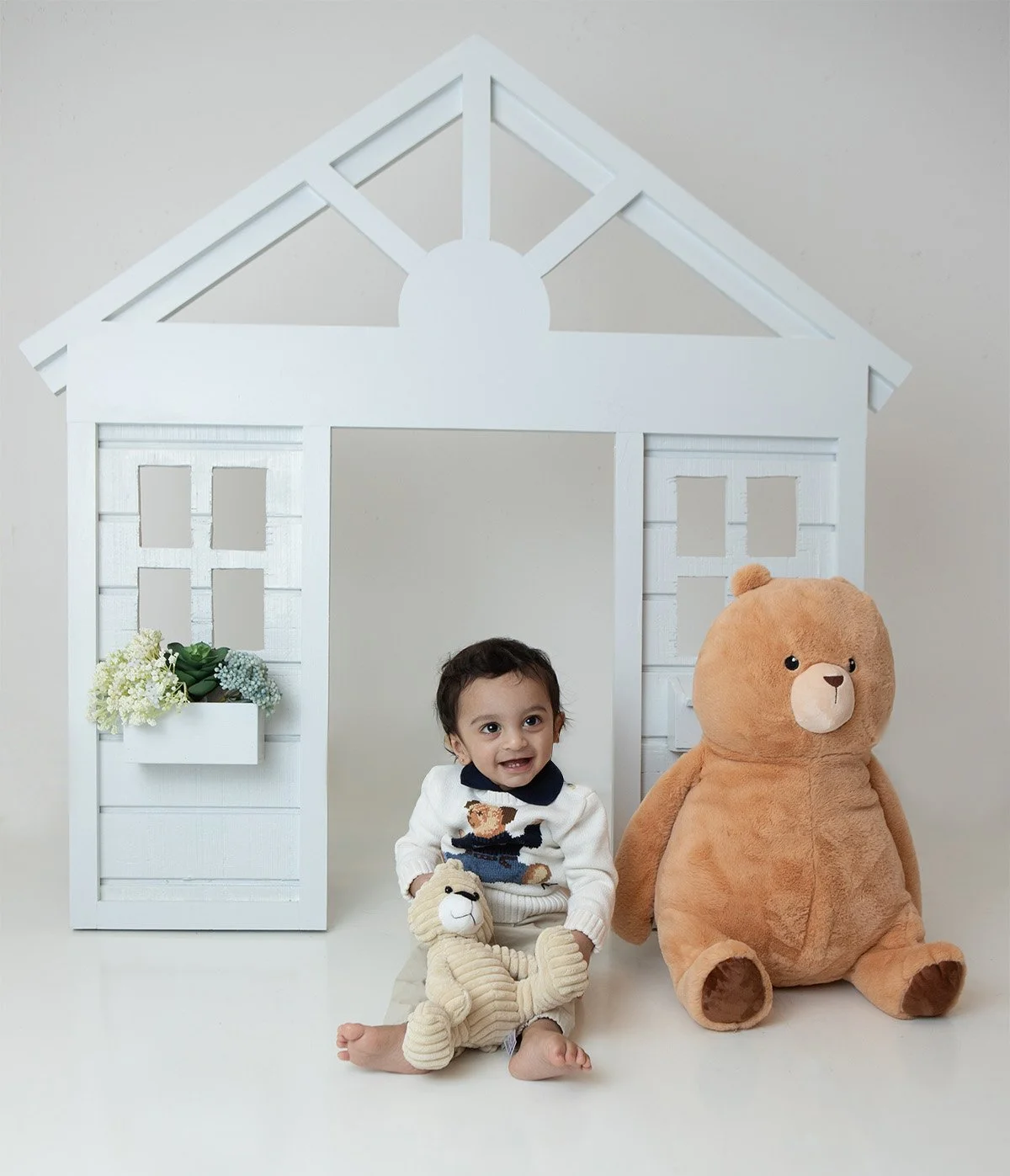 one year old boy sitting ont he floor holding a stuffed bunny with a large stuffed brown bear sitting next to him with a white playhouse 