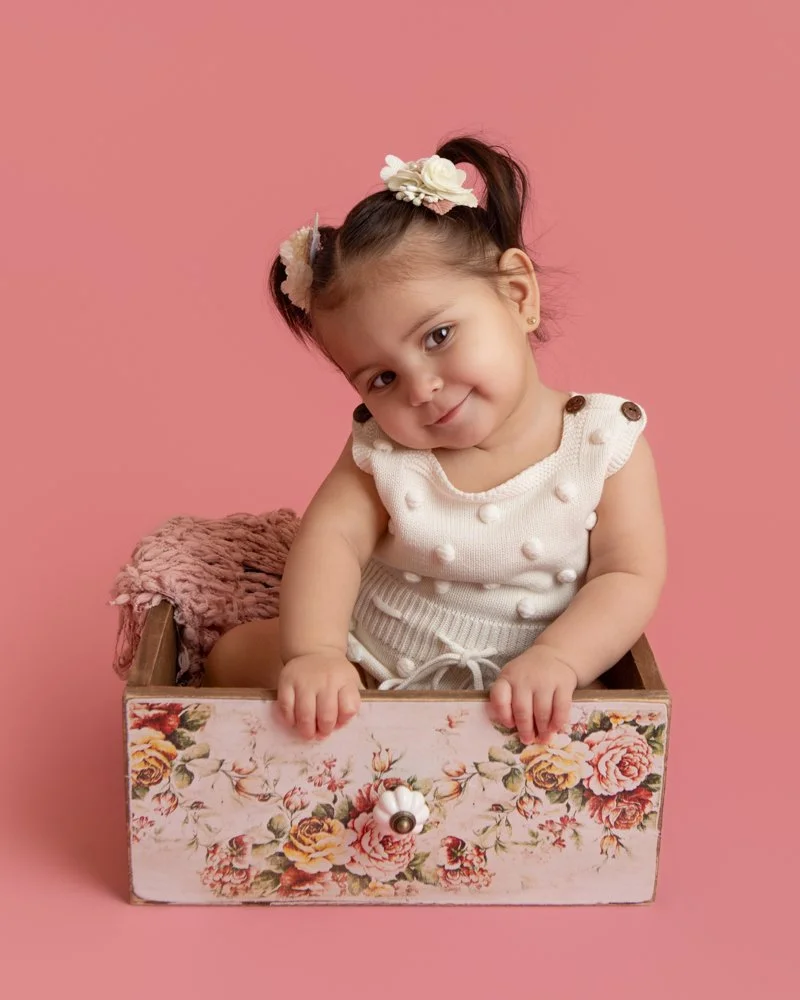 one year old girl sitting in a floral box wearing a white polka dot romper with a pink backrop