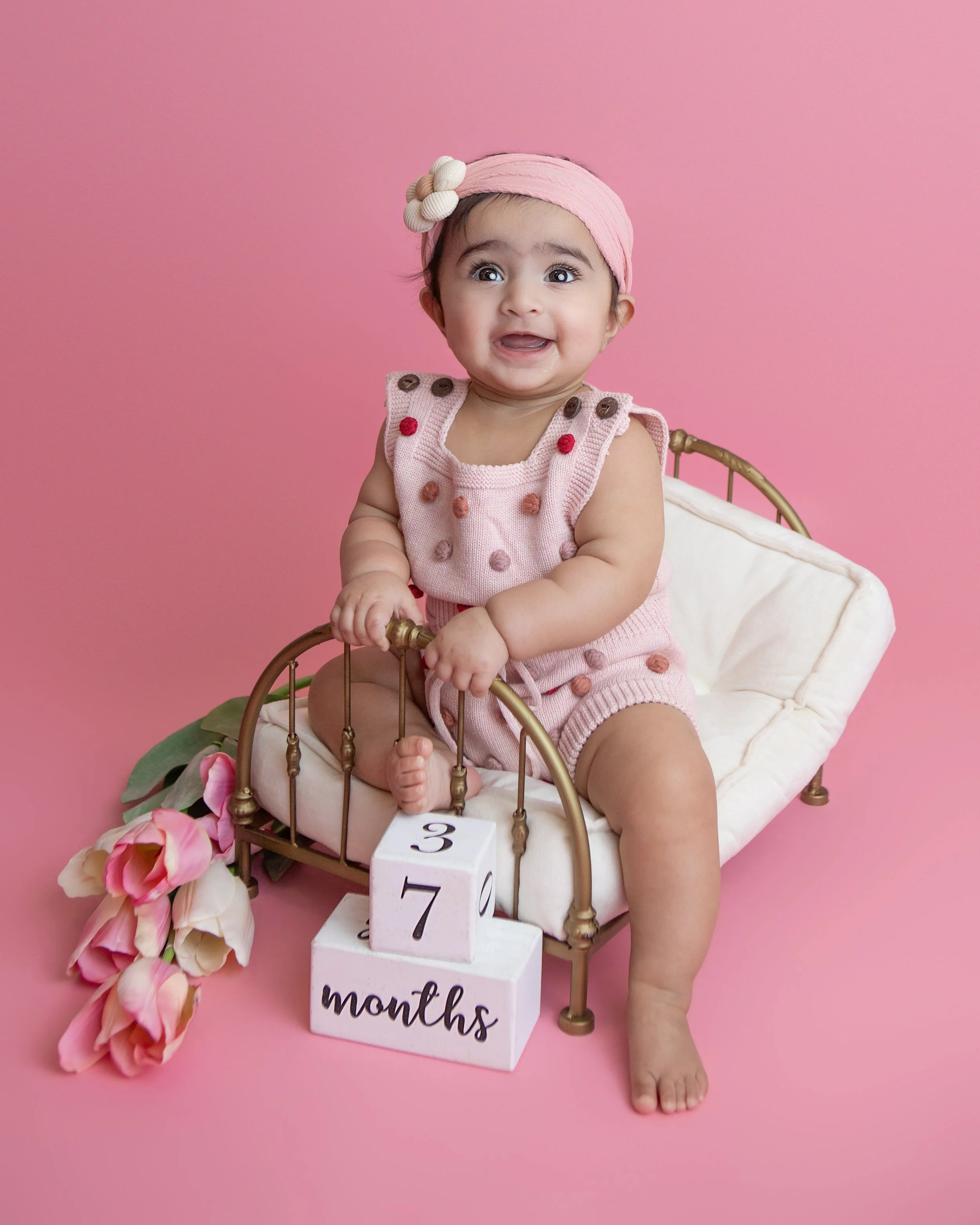 7 month old girl smiling sitting on a brass bad wearing a polka dot romper