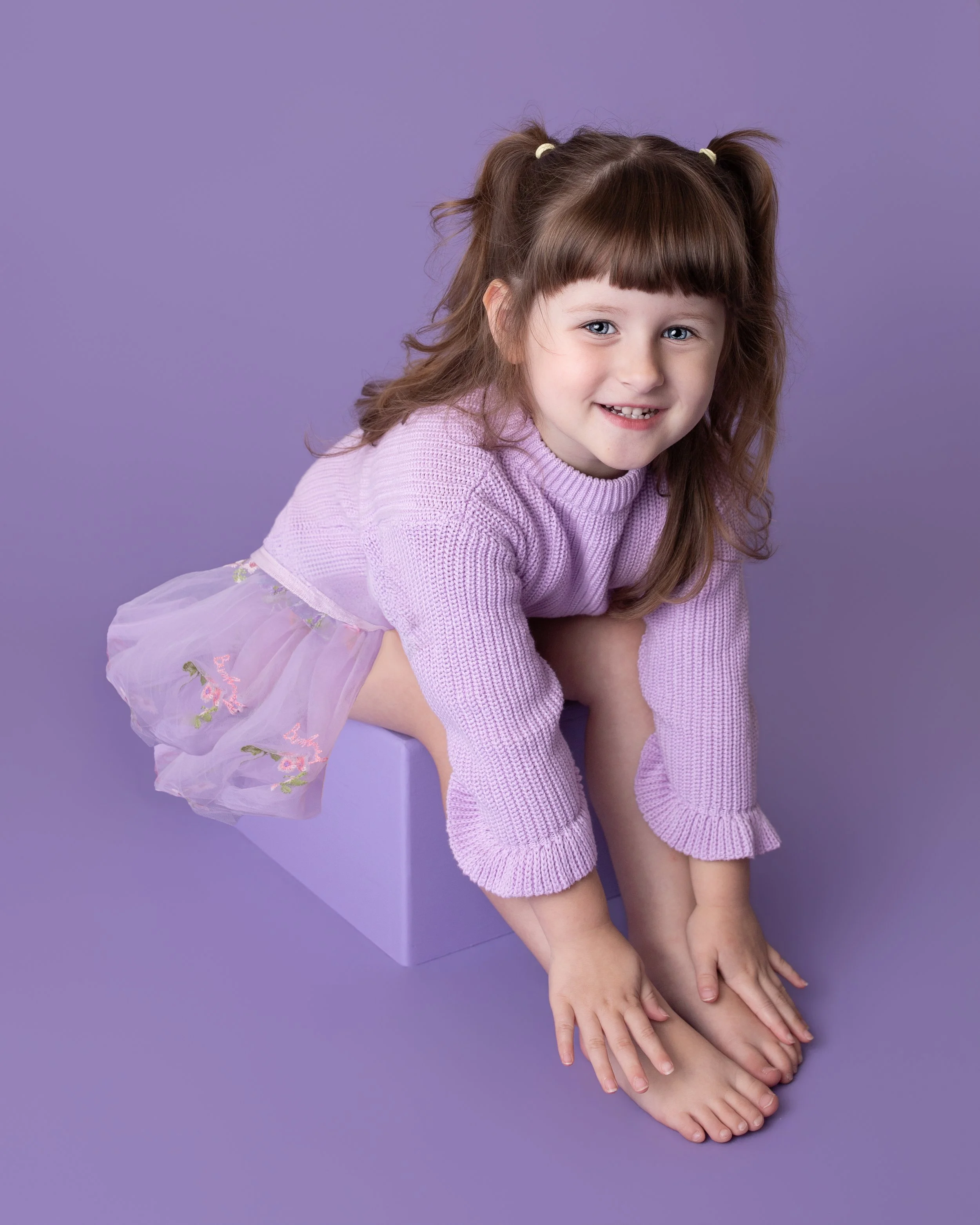 little girl with pony tails leaning over holidng her feet wearing a light purple dress sitting on a light purple box colored box with a light purple colored background
