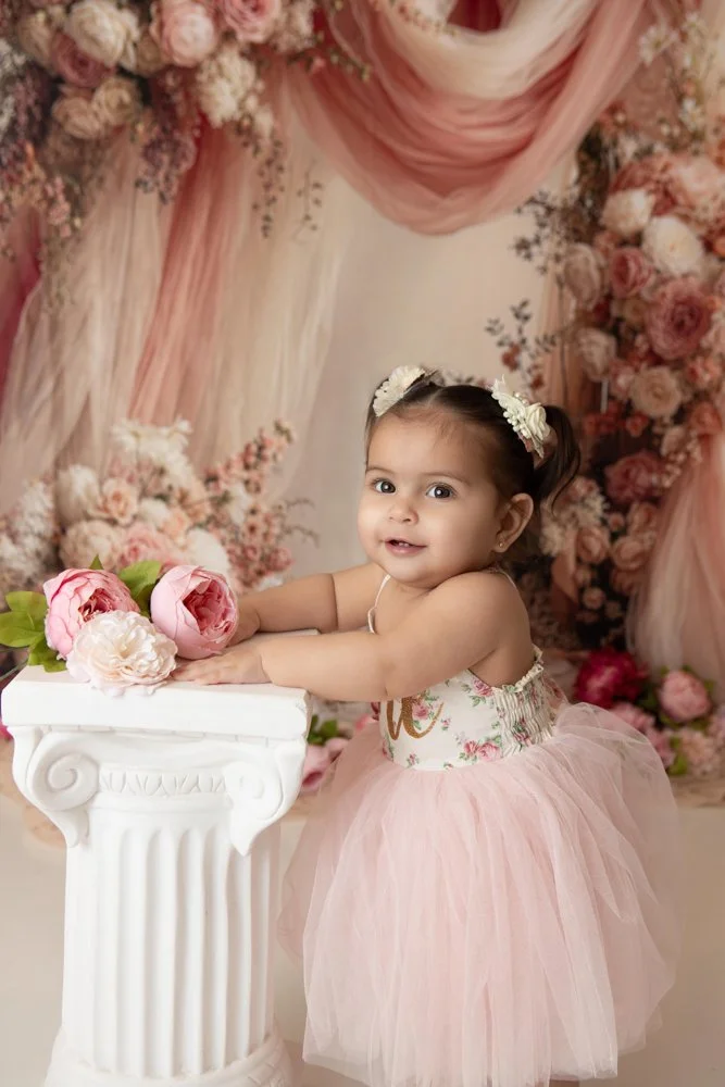 a one year old girl wearing a pink tulle dress holding pink flowers with a pink floral fabric backdrop 