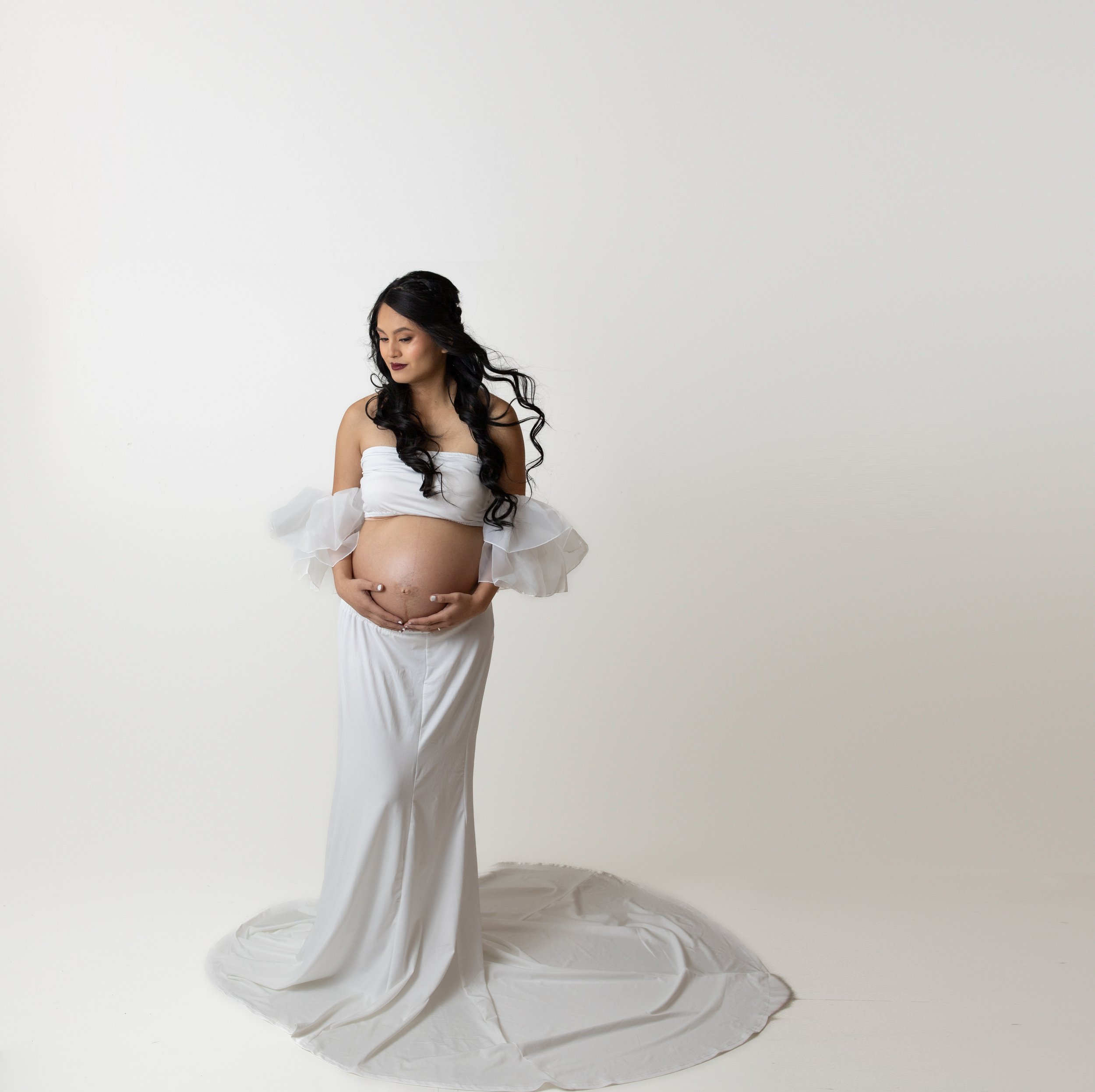 A pregnant woman with long dark hair and makeup, wearing a white off-the-shoulder top with ruffled sleeves and a long white skirt, standing against a plain white background.