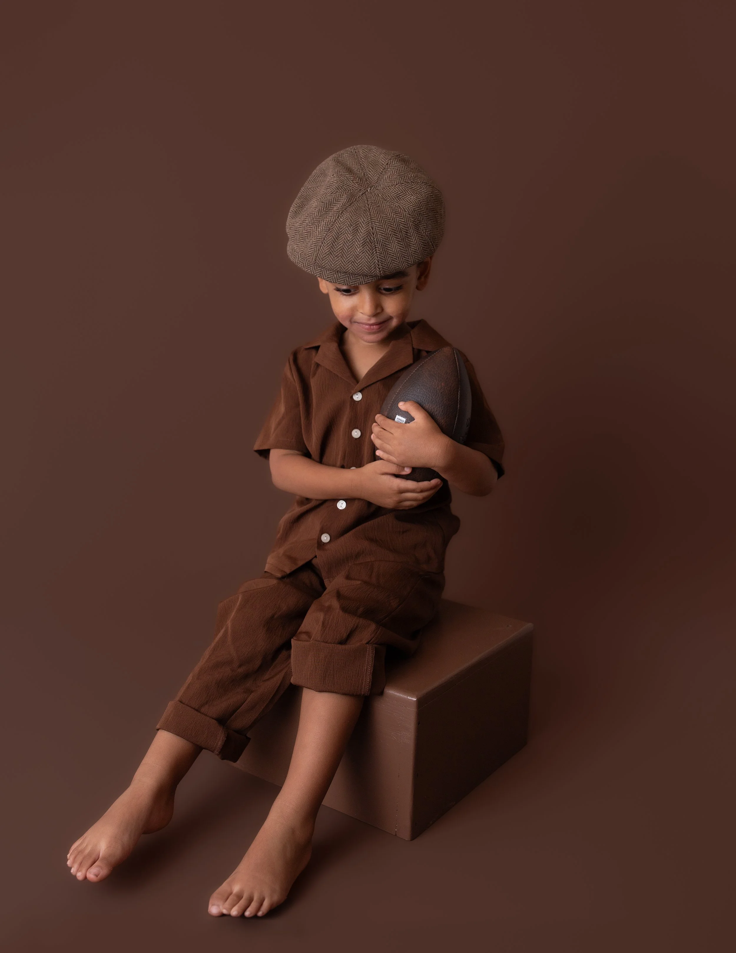 little boy  wearing a tan colored cap and brown outfit holding a football sitting on a brown colored box with a brown colored background