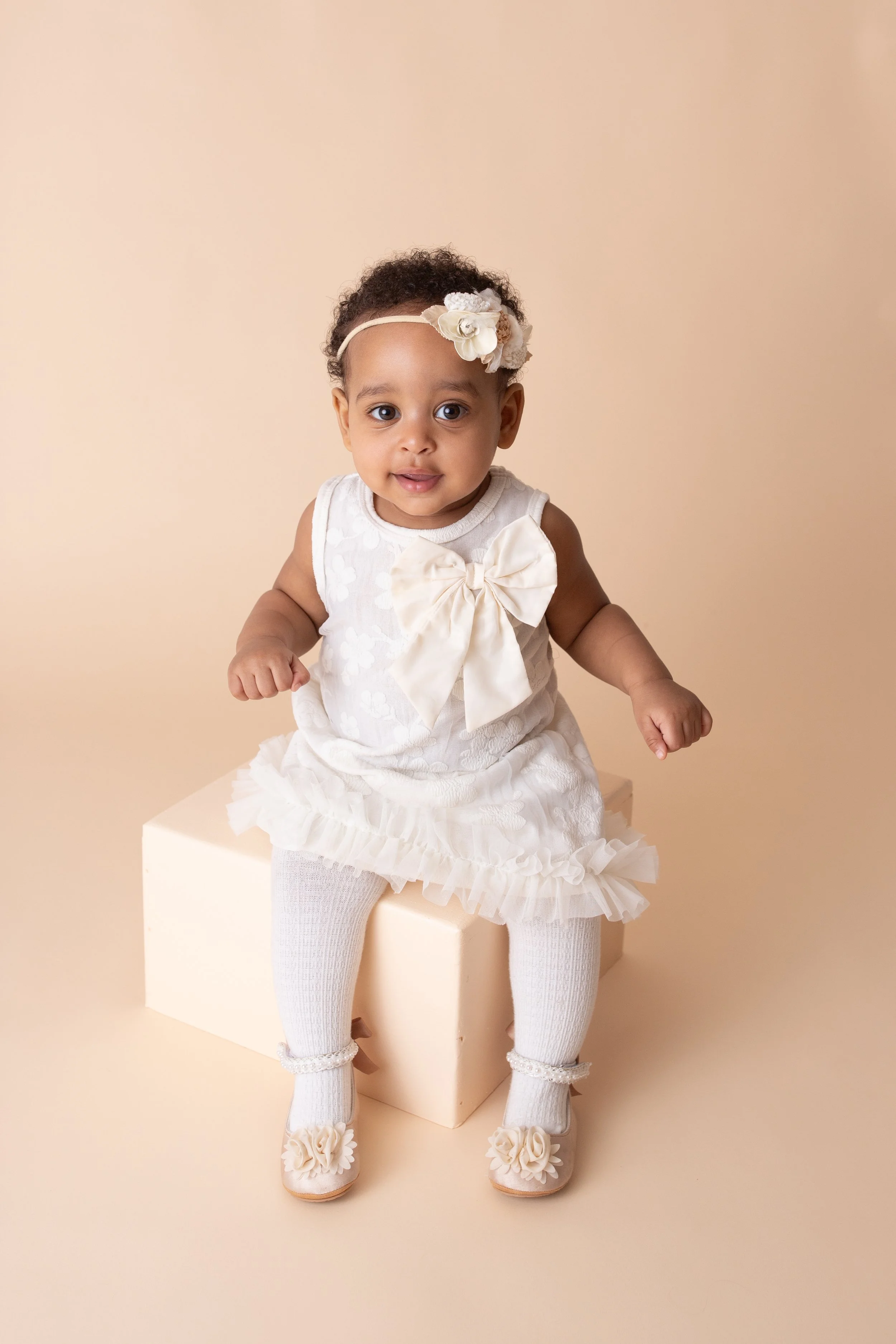 little girl with bow in her hair wearing a cream colored ruffled dress sitting on a cream colored box with a creram colored background
