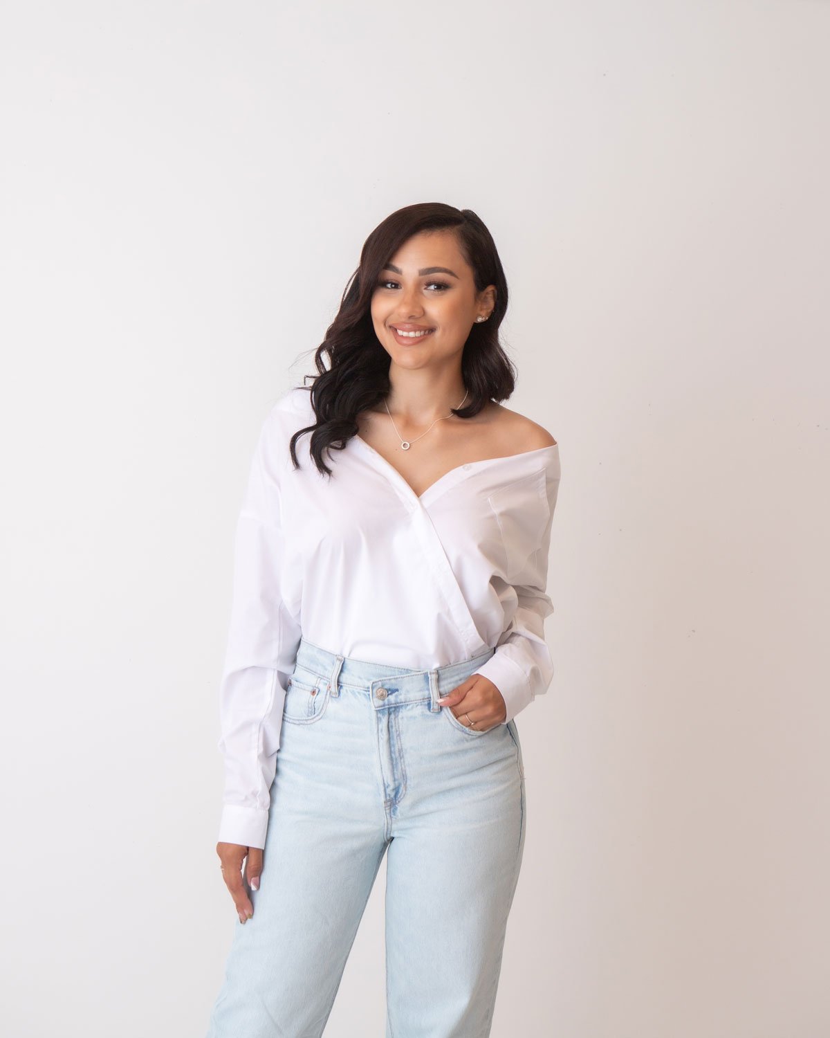 miling young woman in a white off-the-shoulder blouse and light denim jeans, posing confidently against a plain white background. Modern and natural lifestyle portrait photography in Lancaster, PA.