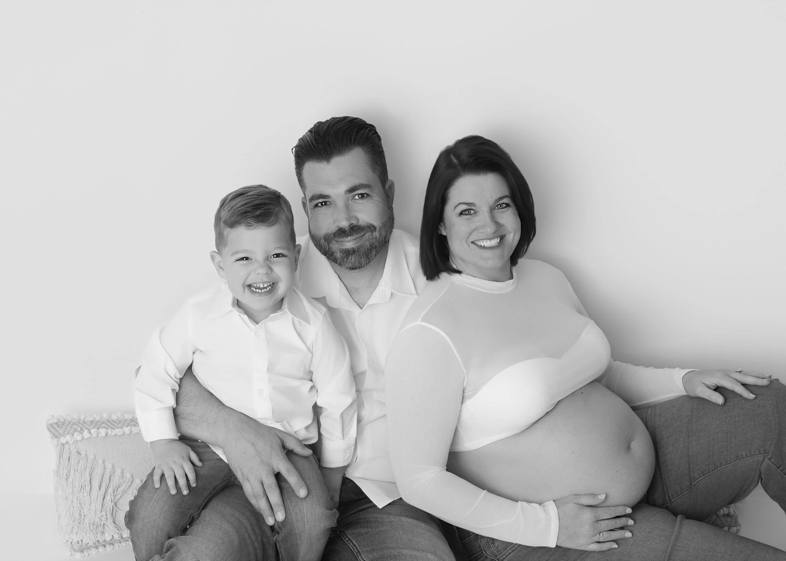 family of three with little boy sitting on floor all smiing and mom is pregant wearing a sheer white crop top