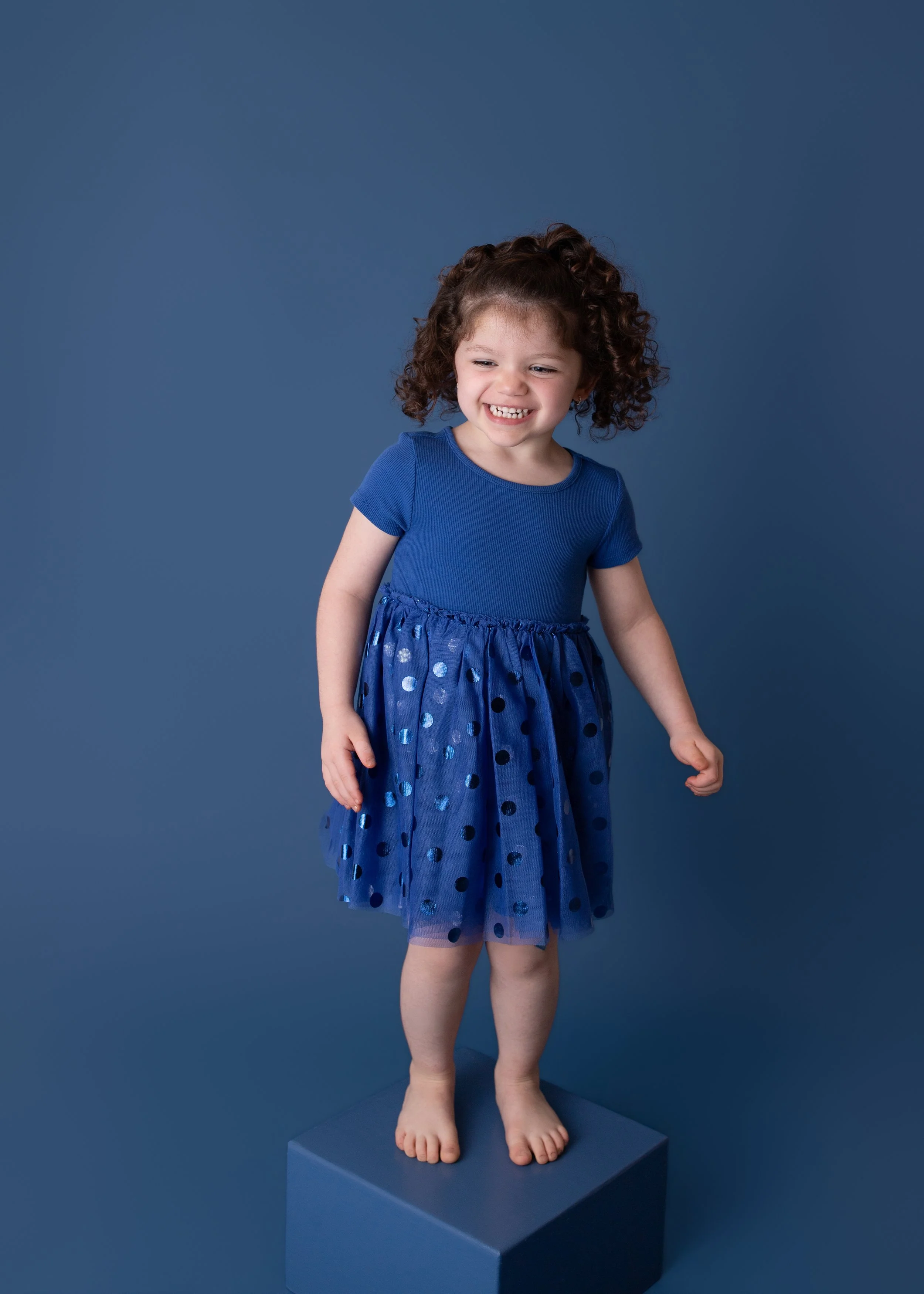 little girl standing on a blue box wearing a blue polka dot colored ruffled dress sitting with a blue colored background