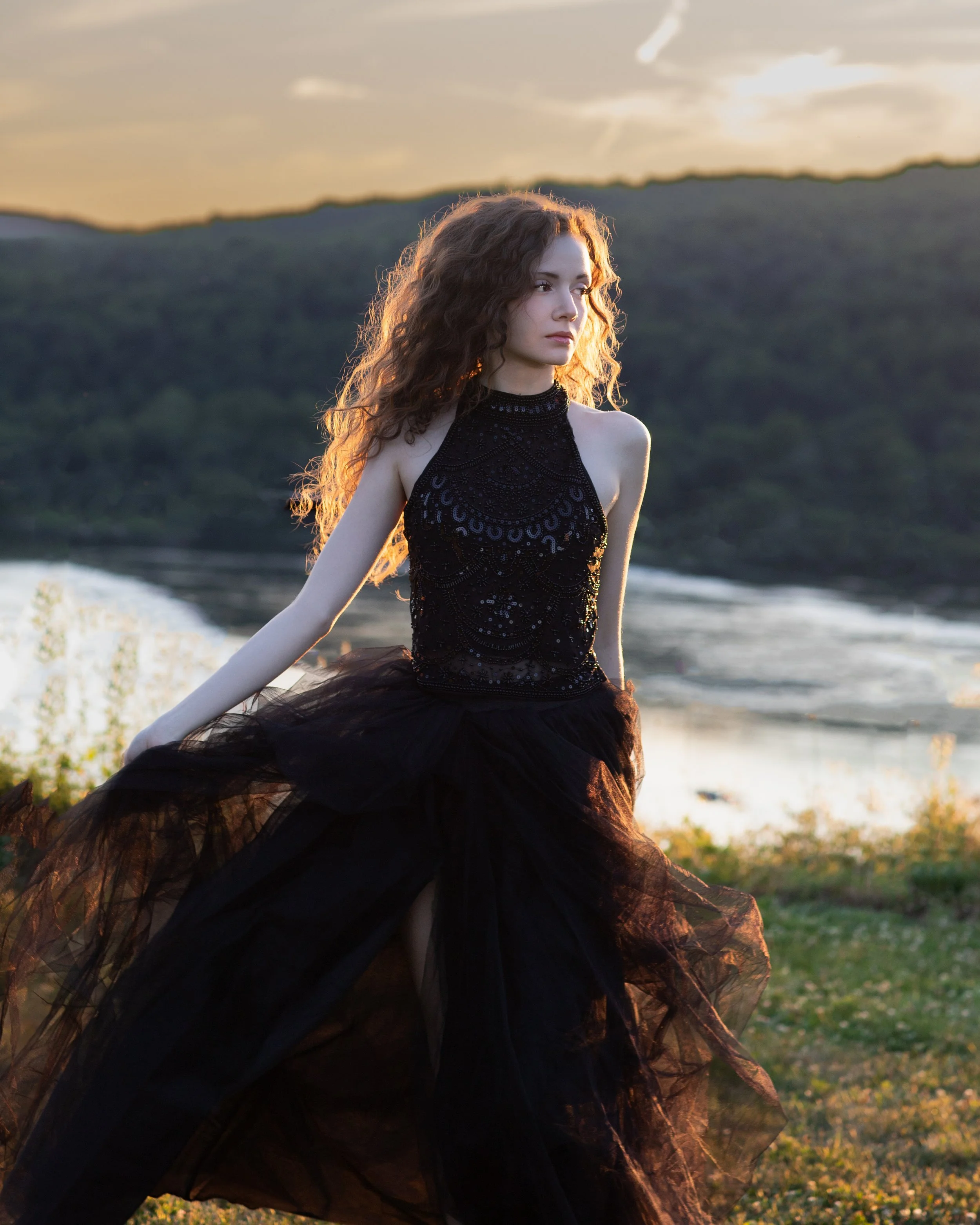 High school senior wearing an elegant black gown posing at sunset along the Susquehanna River in Lancaster, Pennsylvania. Golden backlighting highlights her hair, creating a timeless outdoor senior portrait with a scenic river backdrop.