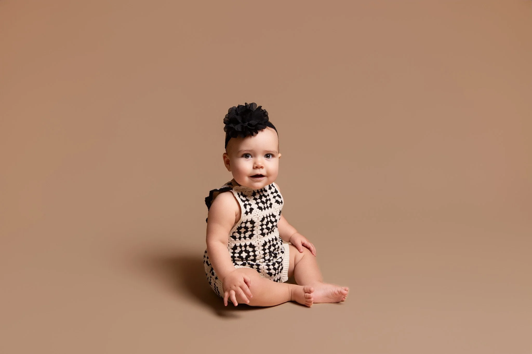 A young baby girl with big eyes sitting on the floor against a plain beige background, wearing a sleeveless black and beige patterned dress and a large black floral headband.