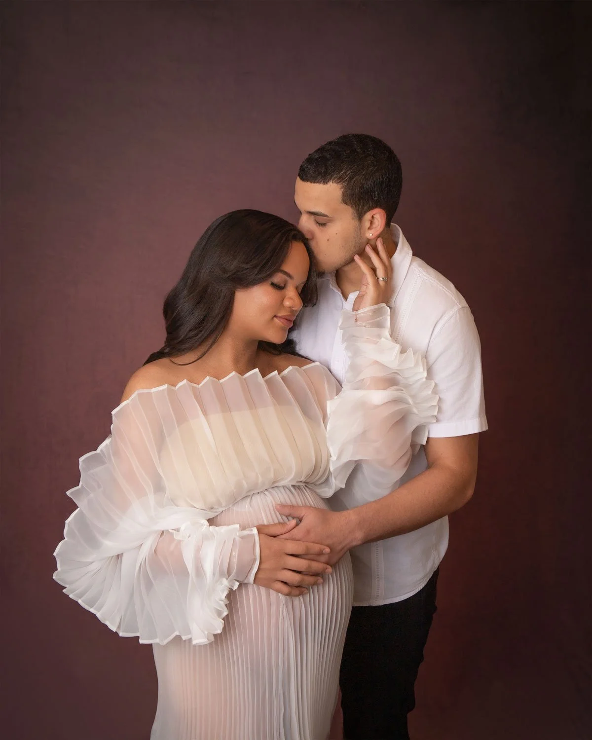 A couple, a woman with dark hair wearing a pleated dress and a man with short hair wearing a white shirt, is embracing each other in a tender moment, with the man kissing the woman's forehead against a plain dark background.