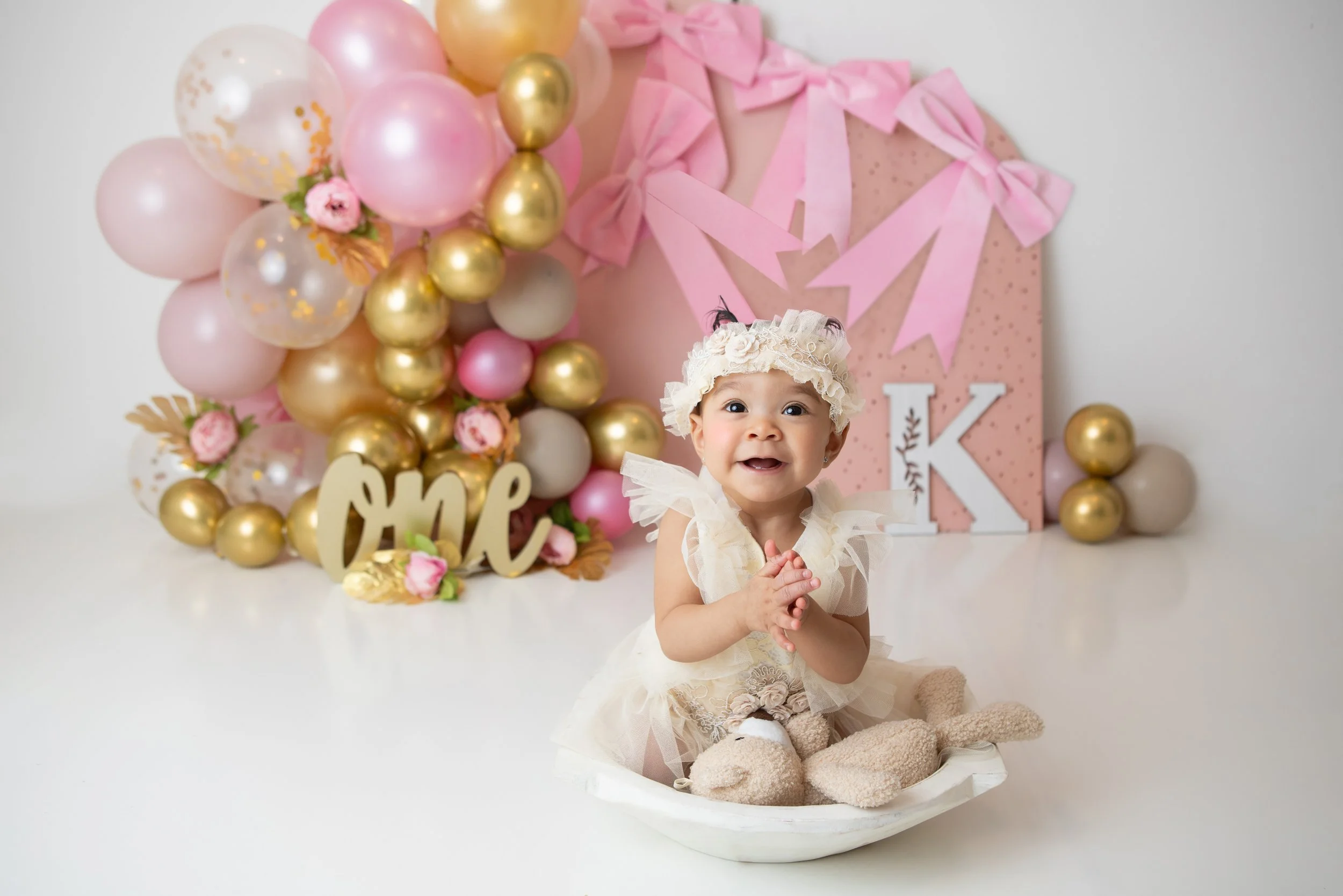 a one year old girl smiling and clapping her hands sitting in a bowl with a pink and gold balloon arch behind her