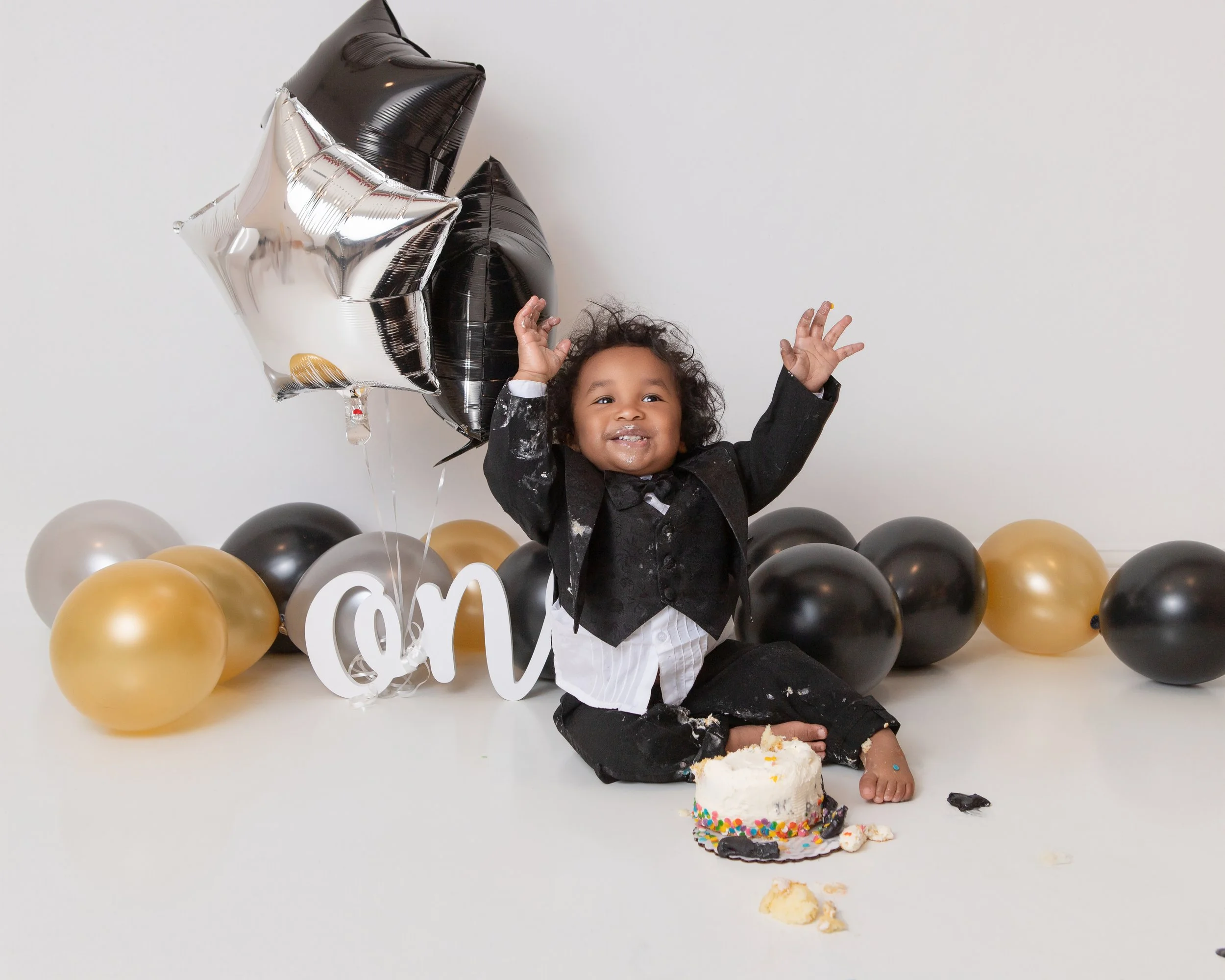 littel boy dressed in a tuxedo with gold silver and black balloons raising his hands with a cake