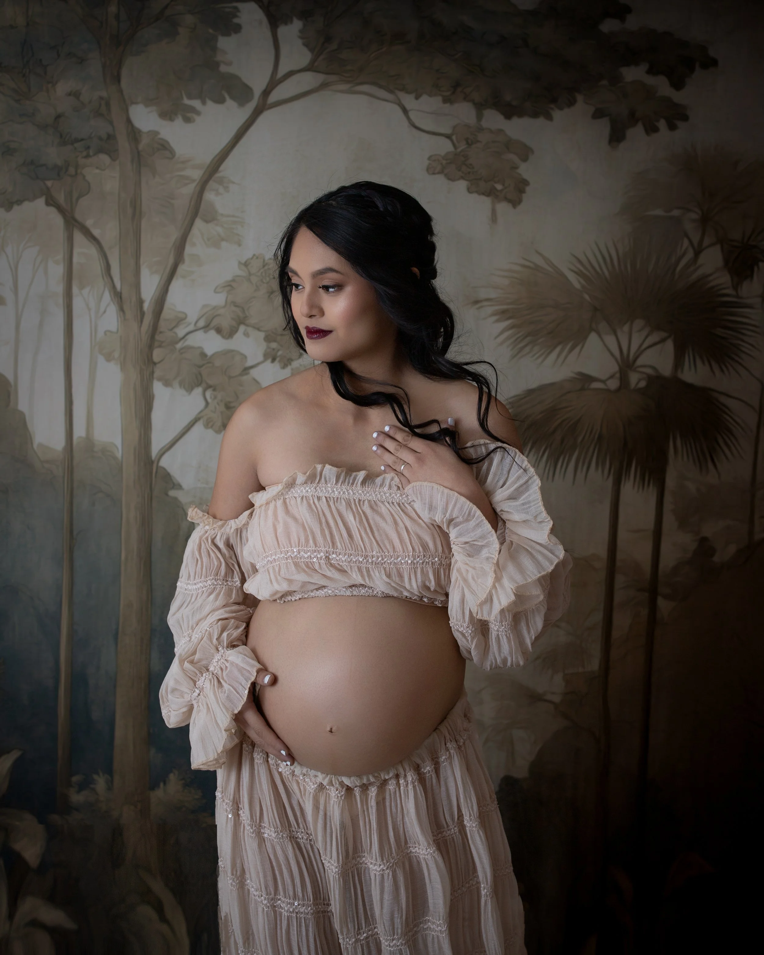 pregnant woman wearing a two piece cream colored dress with long dark hair holding her hand to her chest looking downward with a tree floral background