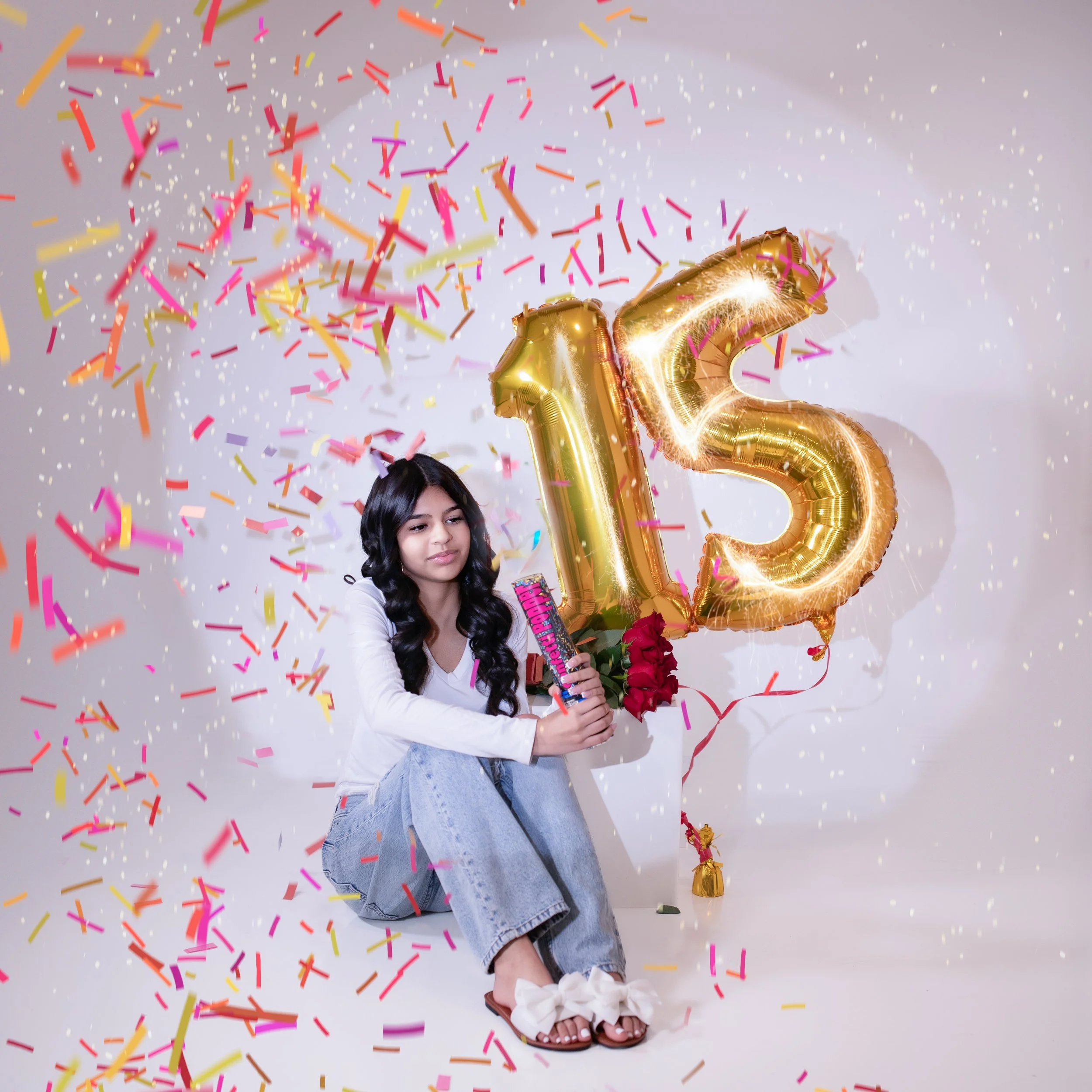 15 year old girl with long dark hair wearing a white shirt with jeans holding te number 15in gold balloons with colorful confetti falling