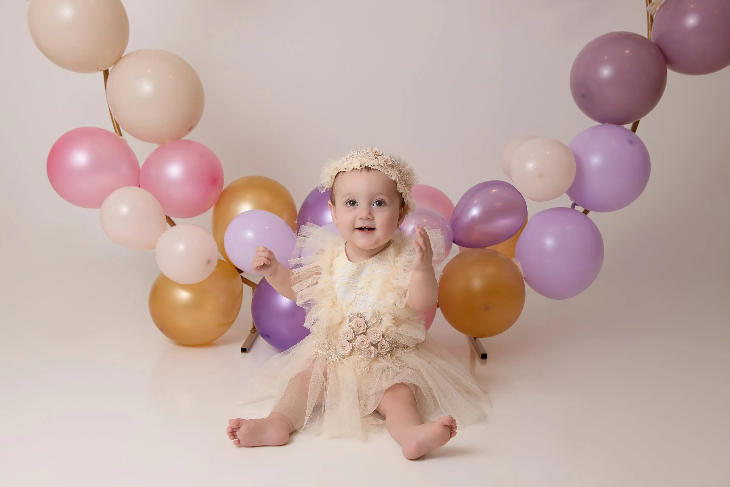 toddler girl first birthday with colorful balloons wearing a cream lacy dress and headband