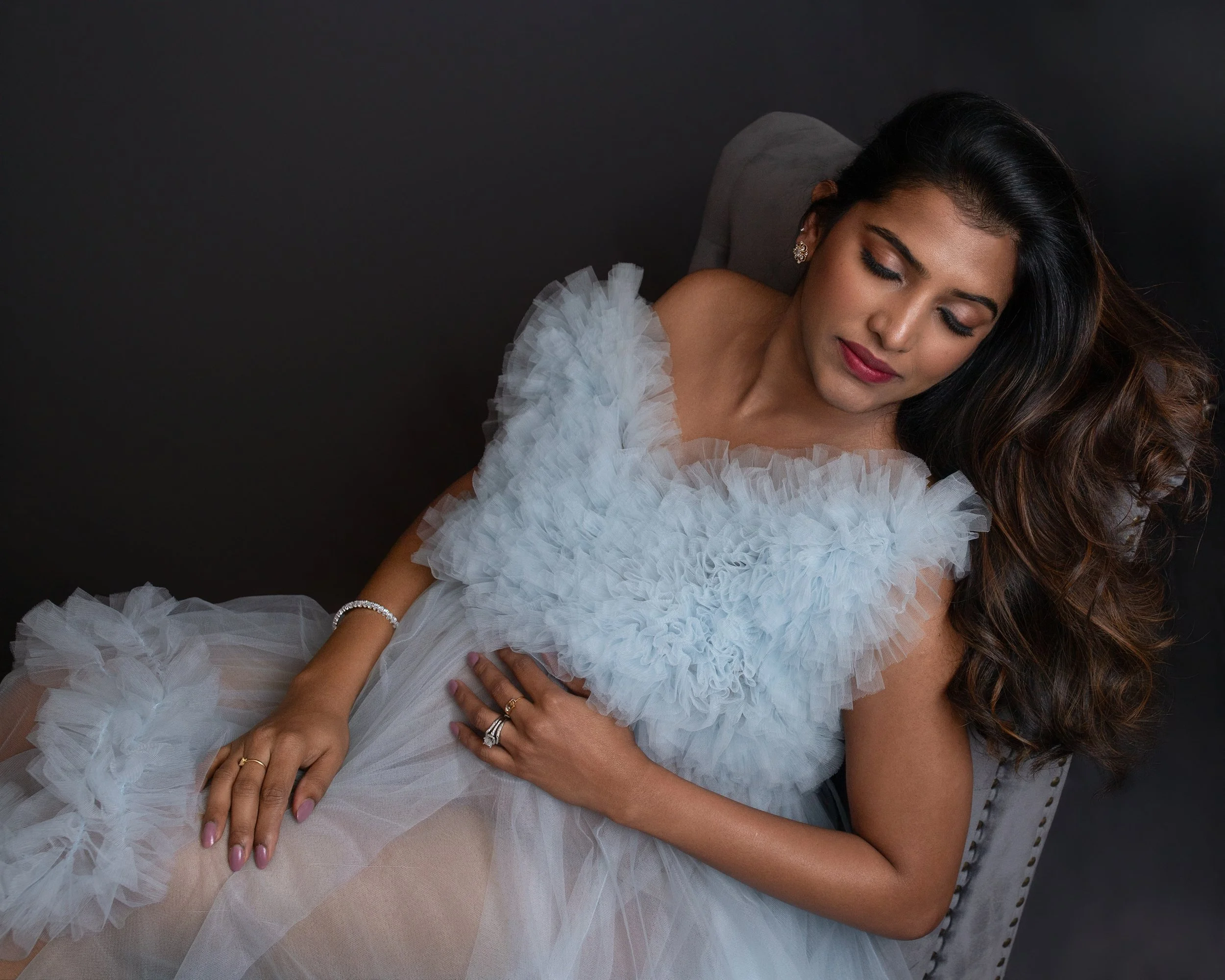 pregnant woman with long brown hair wearing a light blue gown with ruffles eyes closed laying on a chaise loung