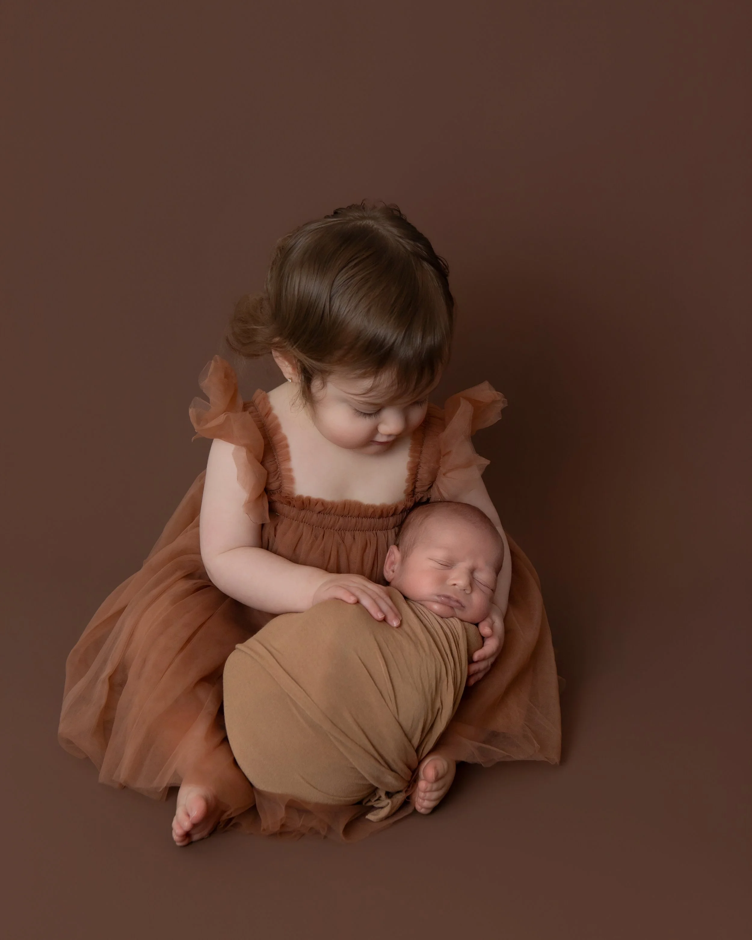 a 2 year old girl wearing a brown dress holidng her newborn brother with a matching swaddle 