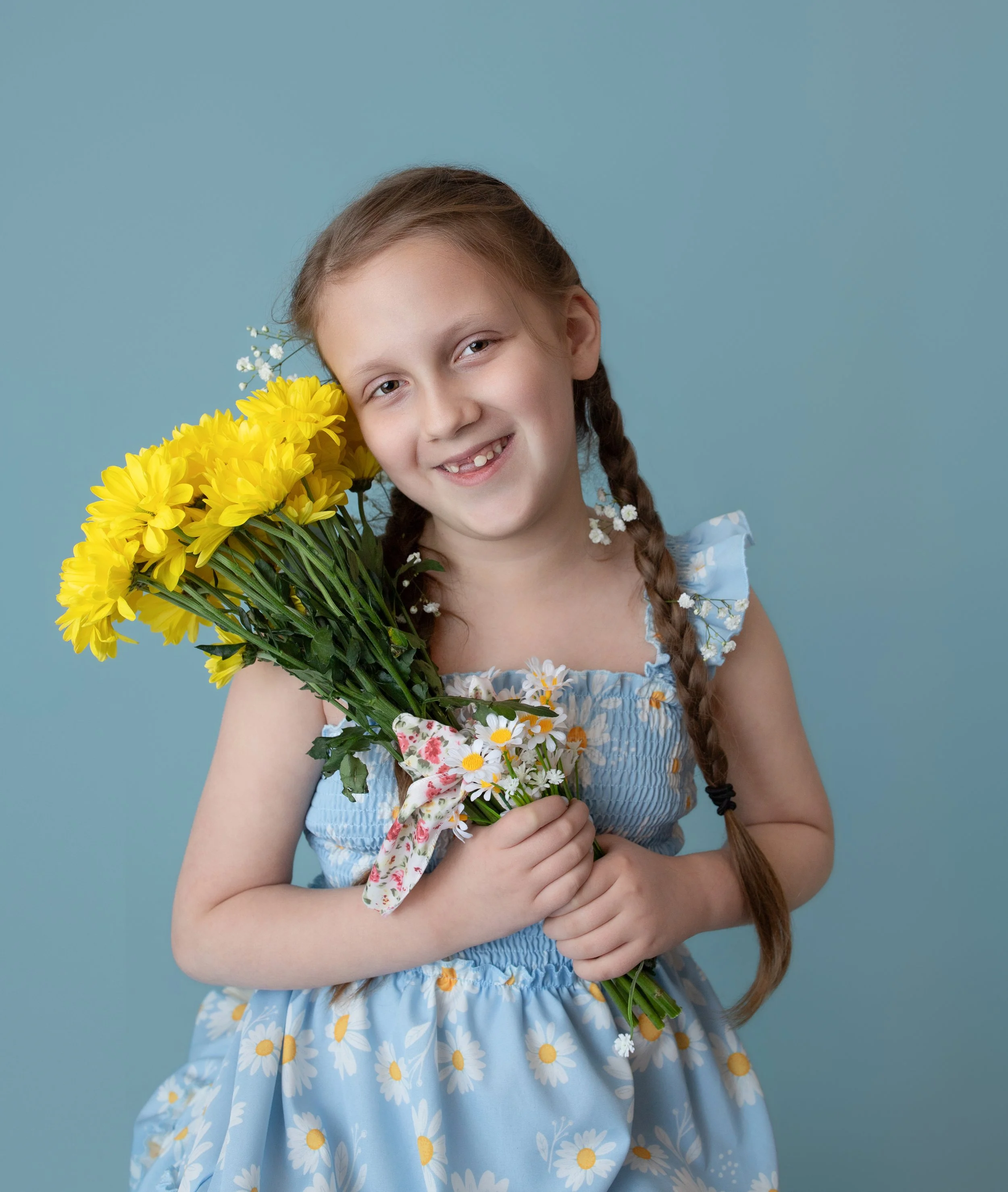 a 5 year old girl smiling holding yellow flowers wearing a light blue dress with yellow flowers with a matching blue backdrop