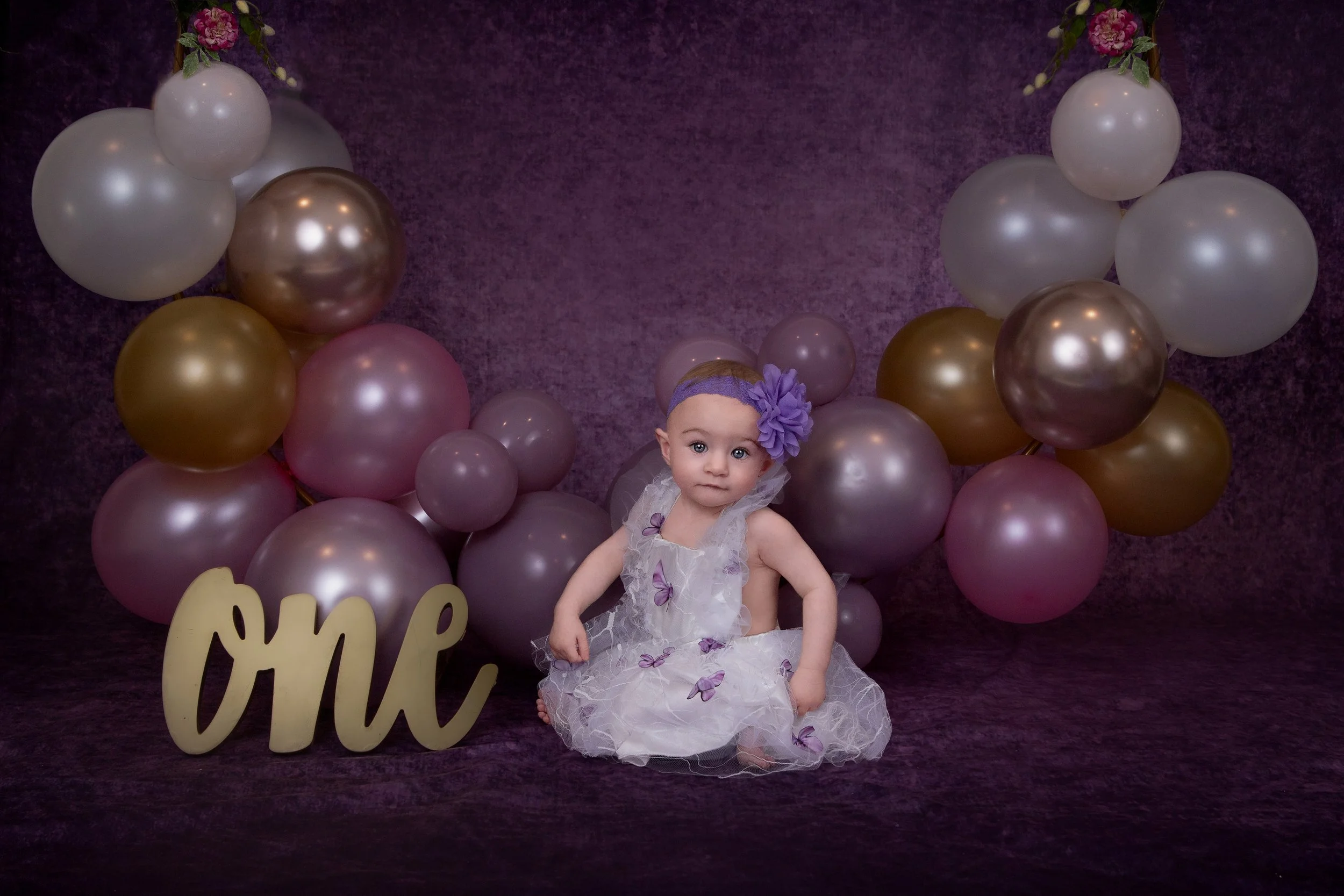 a one year old girl wearing a white dress with purple butterflies with a balloon arch with multi colors