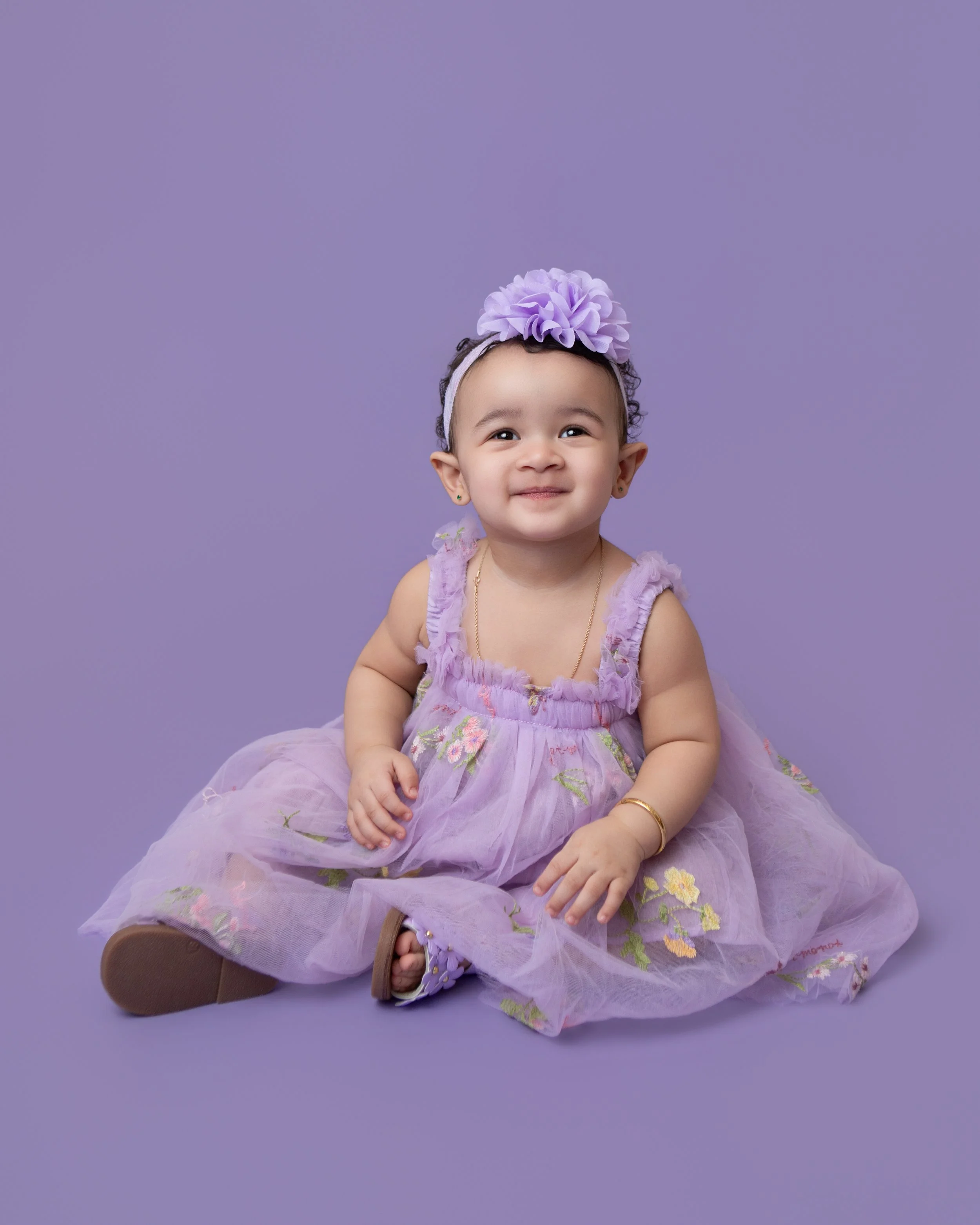 a one year old girl wearing a purple dress and matching headband with a purple backdrop