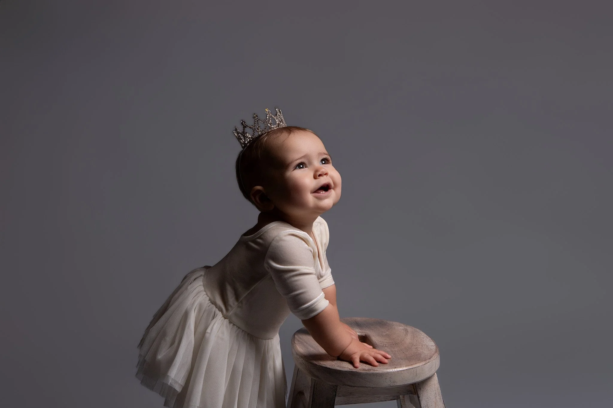 toddler girl wearing a crystal tiara and a velvet cream colored dress with a tutu skirt looking upwards and smiling