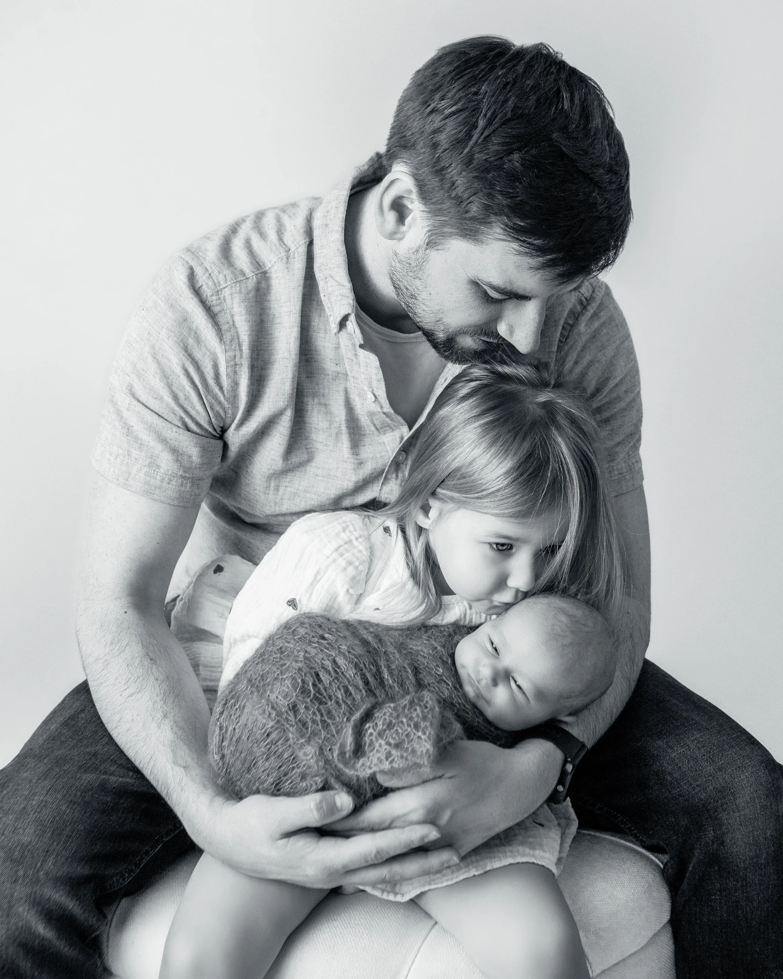 a black and white image of a father holding his toddler and newborn all looking down