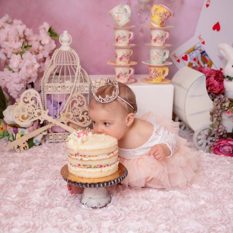 Baby girl dressed as a princess with a tiny tiara, sitting on a pink floral backdrop, curiously examining a layered birthday cake. Elegant and whimsical cake smash photography in Lancaster, PA.