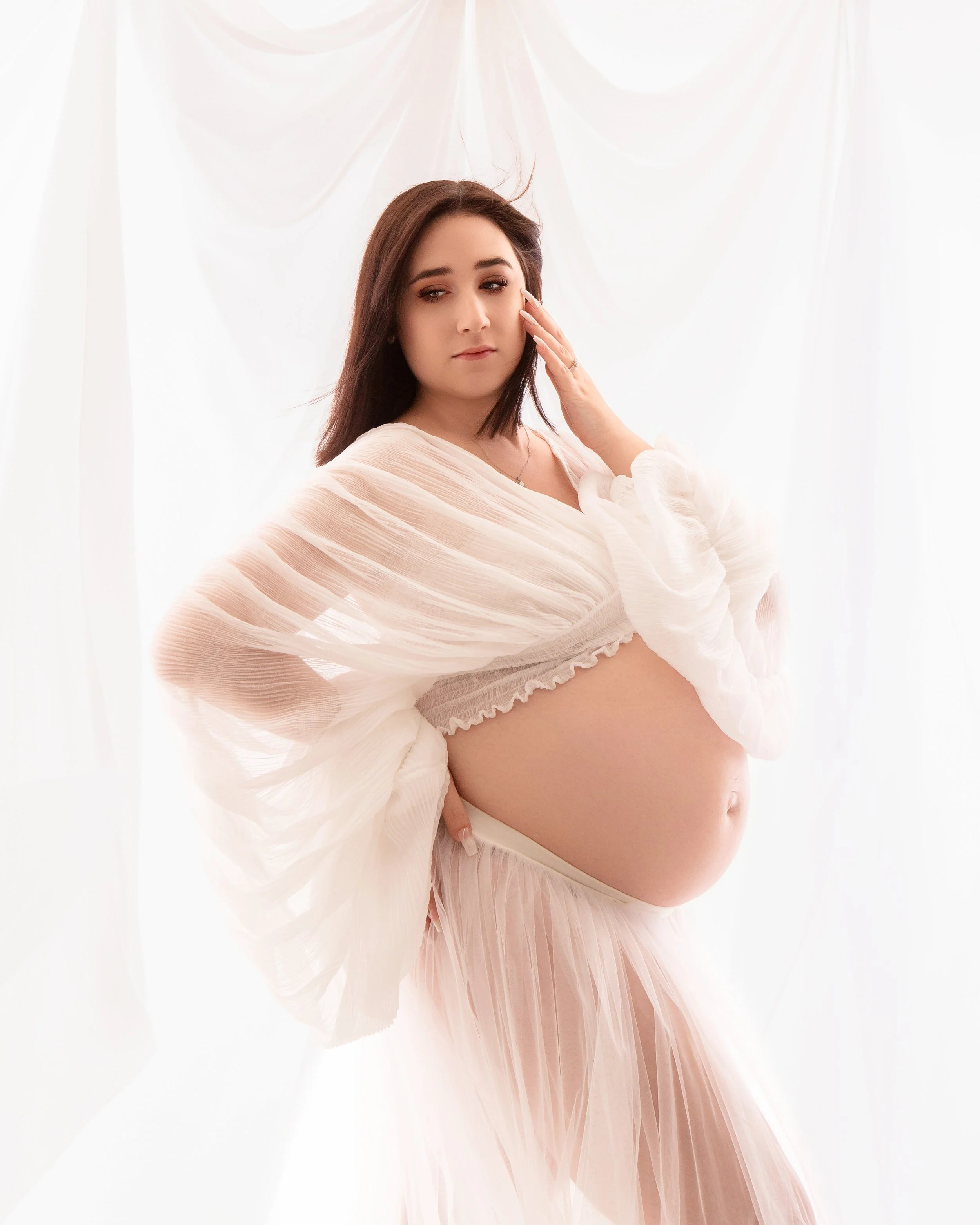 a pregnant women wearing a white sheer top with a white sheer skirt holdingher hand to her cheek