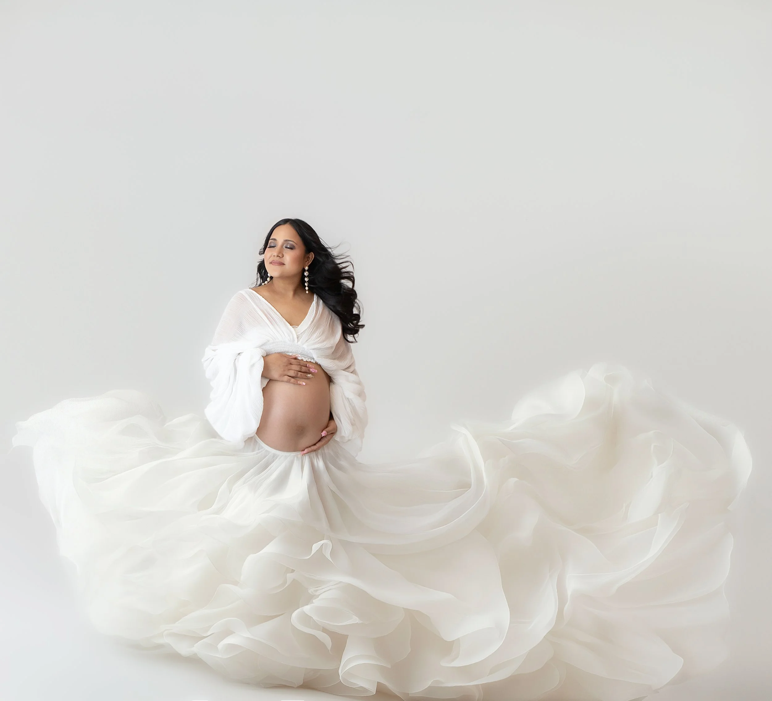 woman wearing a white flowing gown with her hair blowing as she looks up holidng her pregnant belly