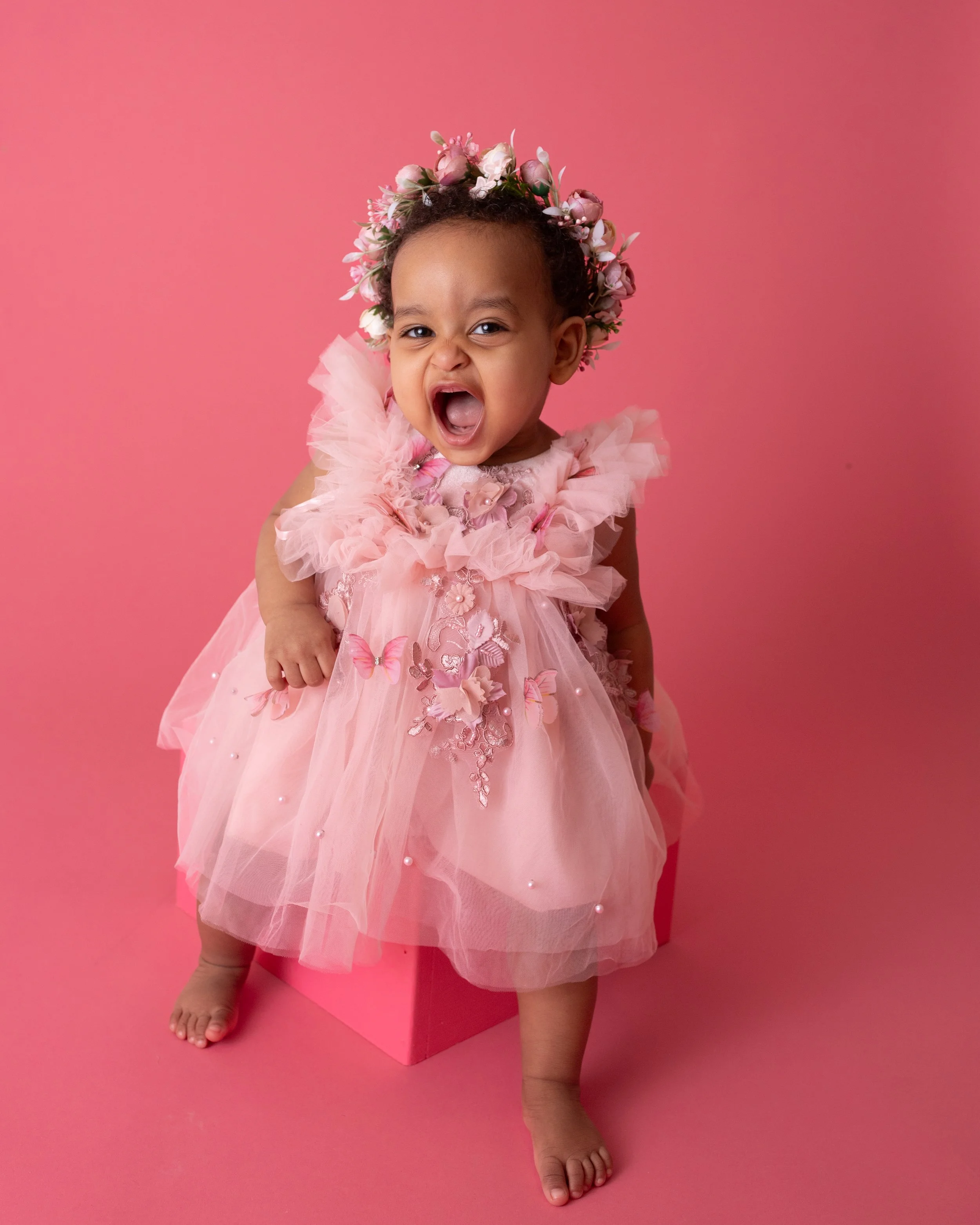 little girl with a big open smile floral wreath in her hair wearing a pink colored ruffled dress sitting on a pink colored box with a pink colored background
