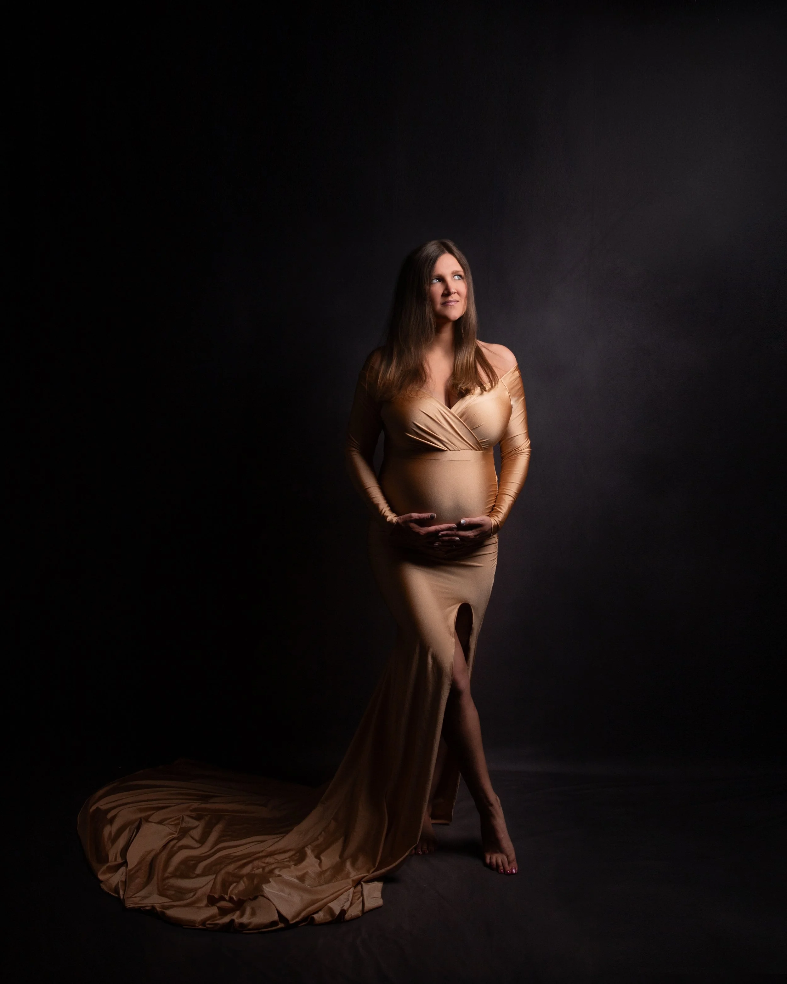 a pregnant women holding her stomach with both hands wearing a gold gown looking up towards the light