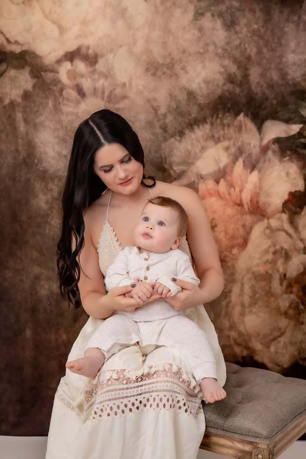 Mother in a cream dress holding her baby, sitting against a vintage floral backdrop, gazing down lovingly. Warm and timeless motherhood portrait photography in Lancaster, PA