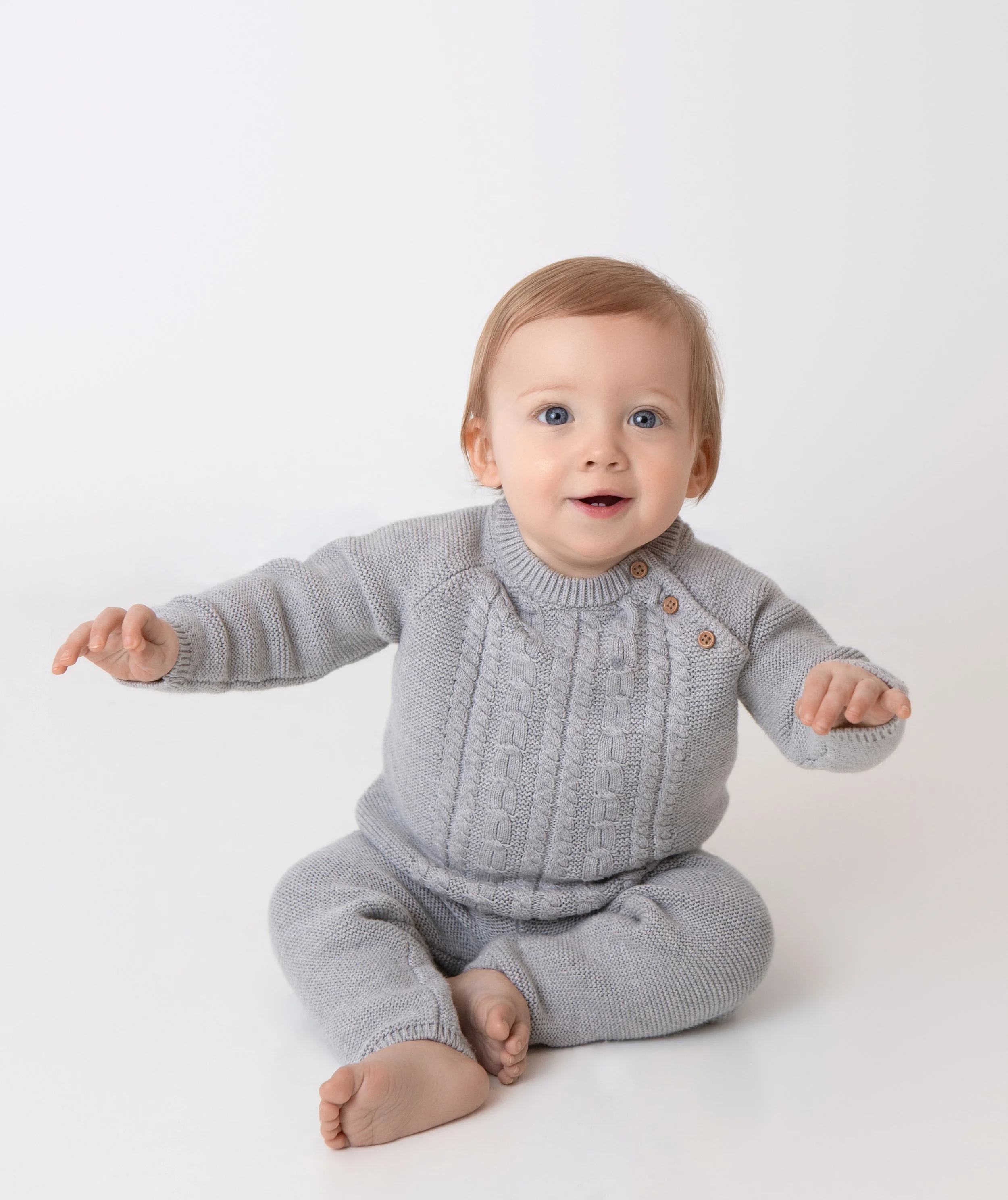 a smiling one year old boy holding both arms up on a white background
