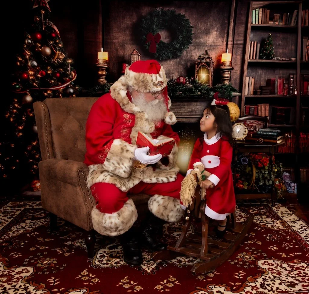 Santa Claus reading a book to a young girl dressed in a Christmas outfit in a festive room decorated with a Christmas tree, wreath, and ornaments.