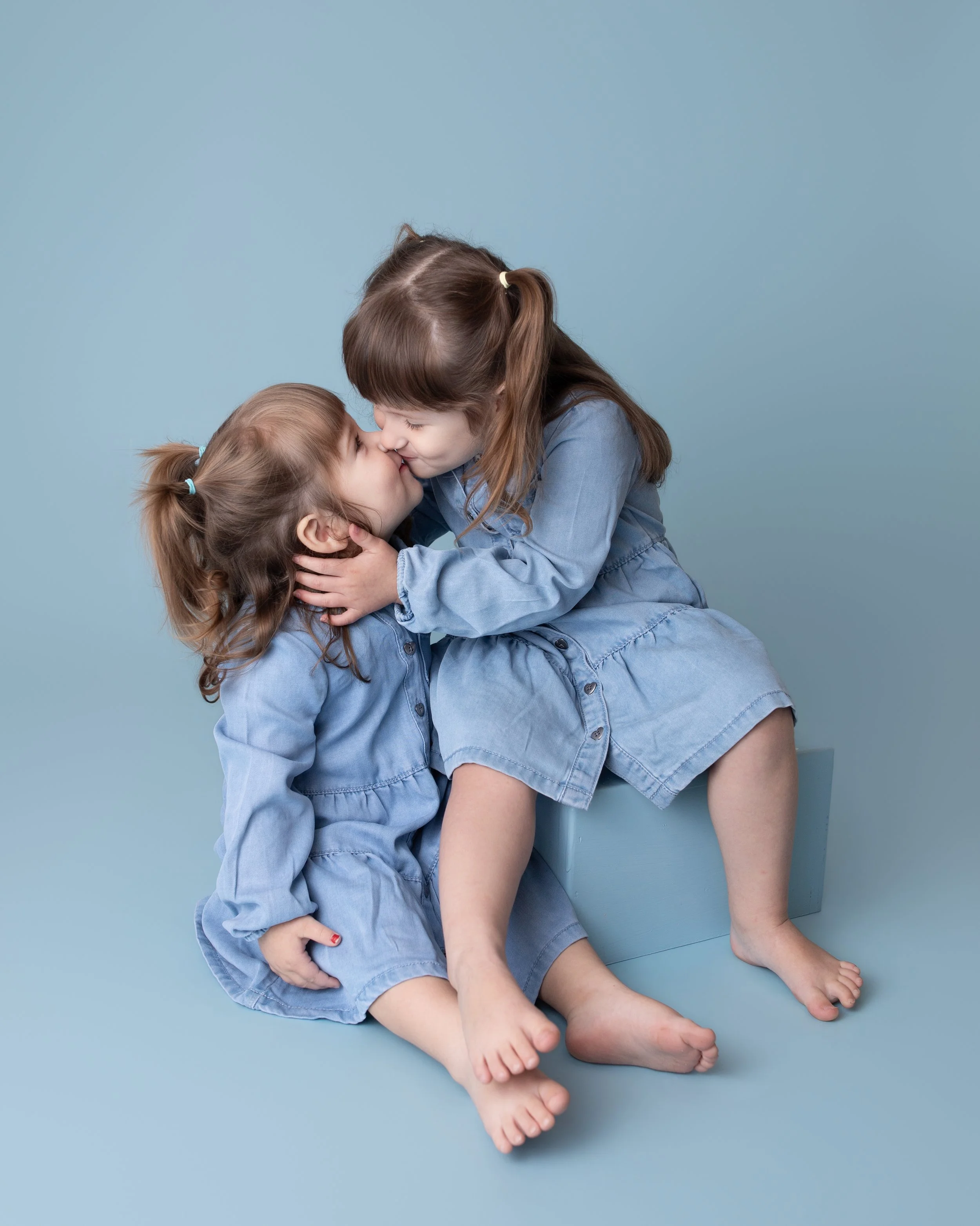 two little girl wearing matching denim dresses giving eachother a kiss sitting on a light blue colored box with a light blue colored background