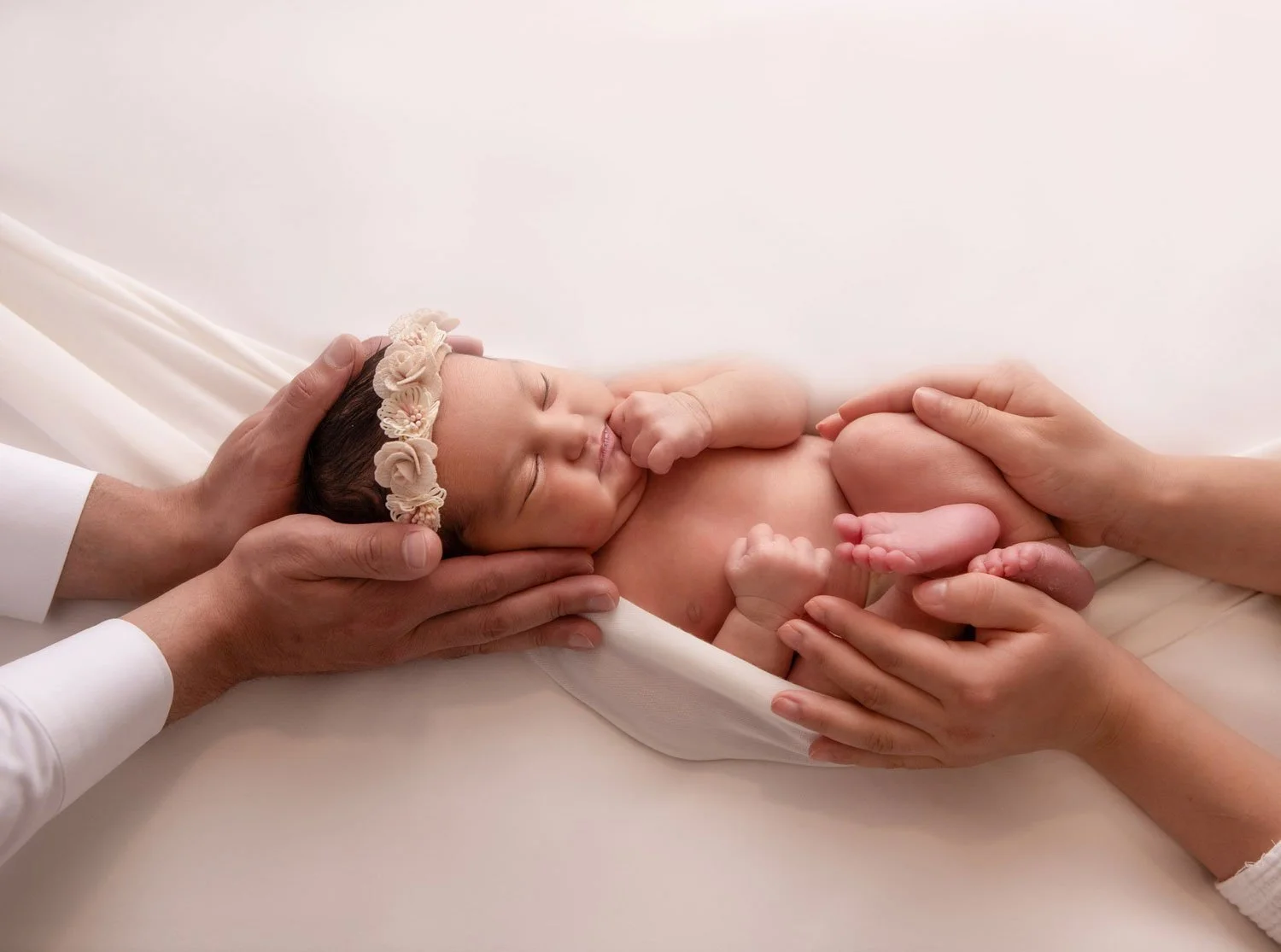 Newborn baby peacefully sleeping, cradled in the loving hands of parents, wearing a delicate floral headband. Heartfelt and intimate newborn photography in Lancaster, PA.