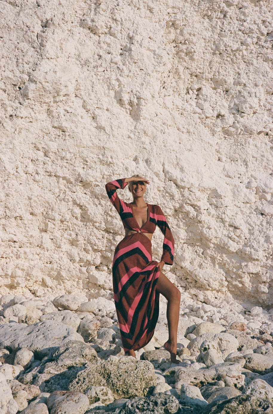 Person in a striped dress standing on rocky beach against a textured cliff face.
