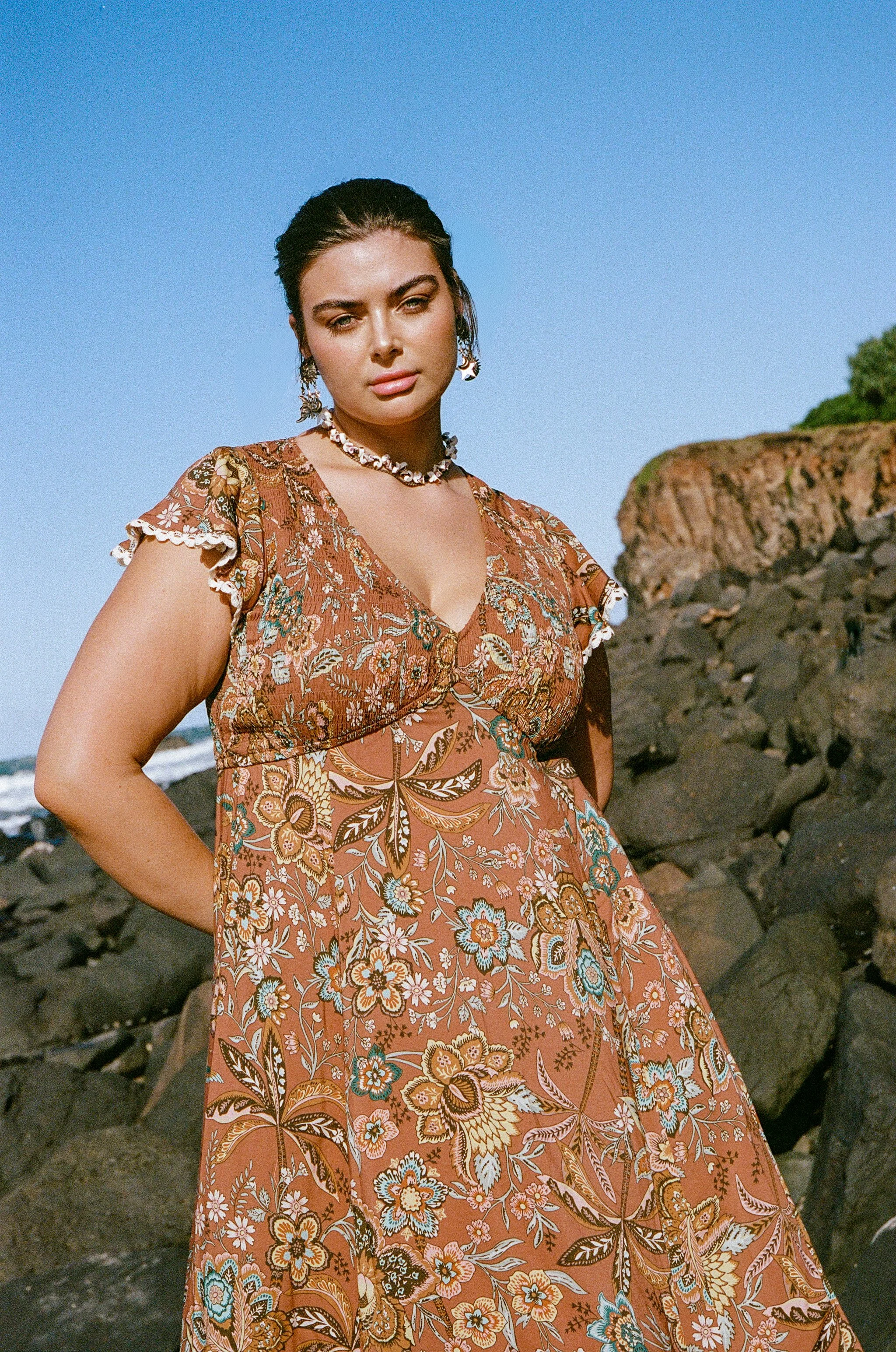 Woman in a floral dress standing on rocky coastline against a clear blue sky.