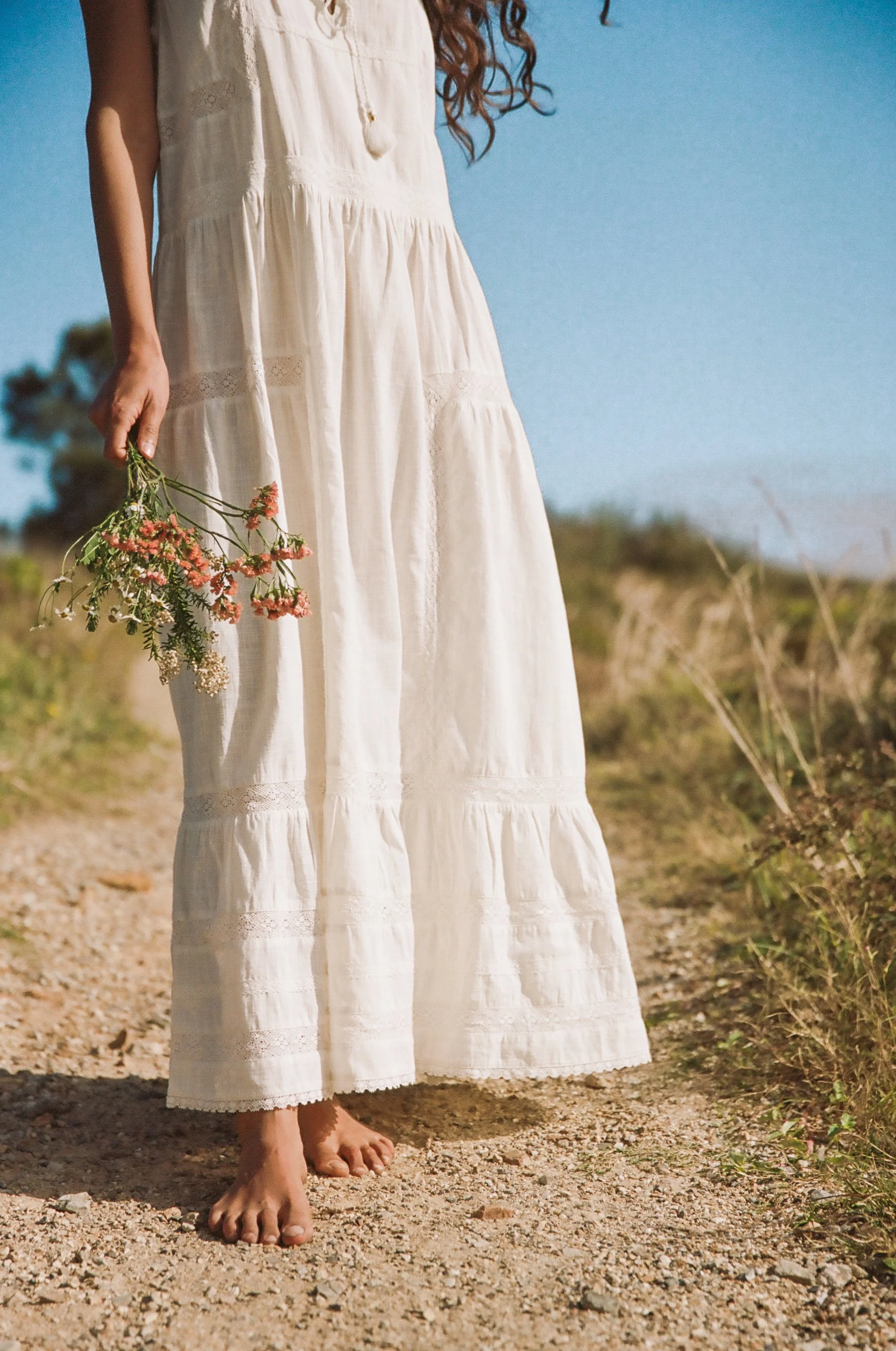 Person in white dress holding wildflowers, standing barefoot on a dirt path.