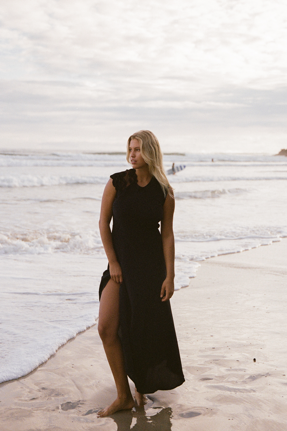 Woman in a black dress standing on a beach with waves in the background.