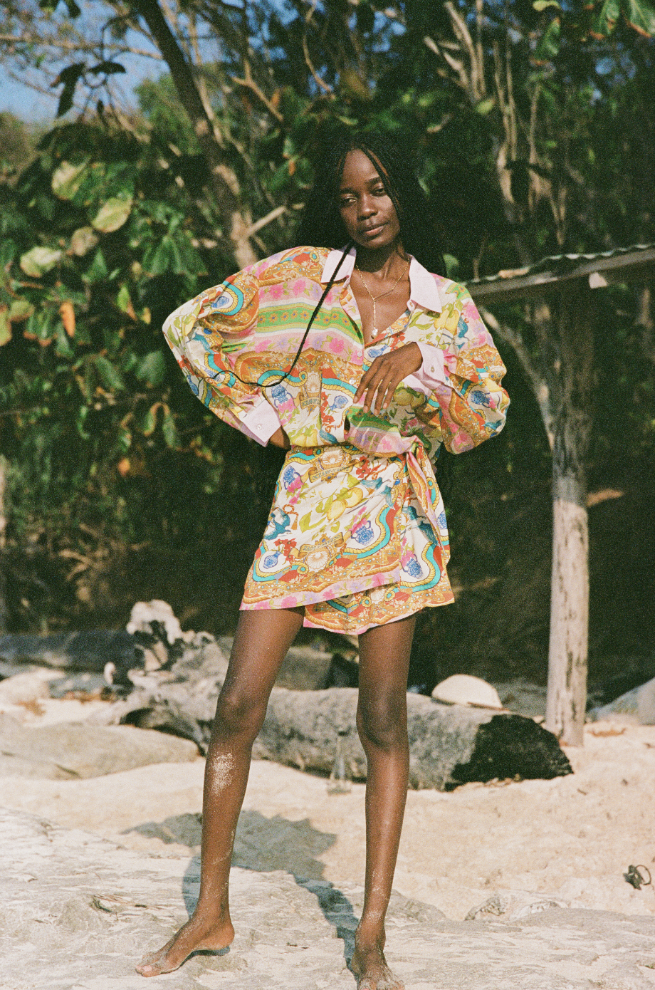 Woman in colorful dress standing outdoors on rocky ground with trees in the background.