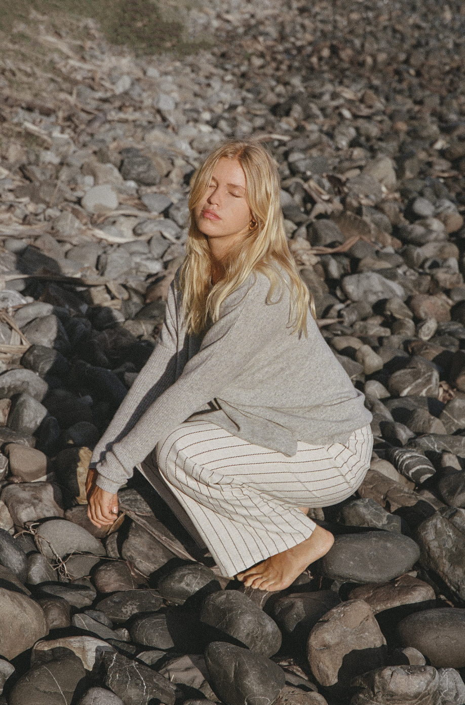 Person with long blonde hair in a gray sweater and striped pants, kneeling on rocky ground, eyes closed, under sunlight.